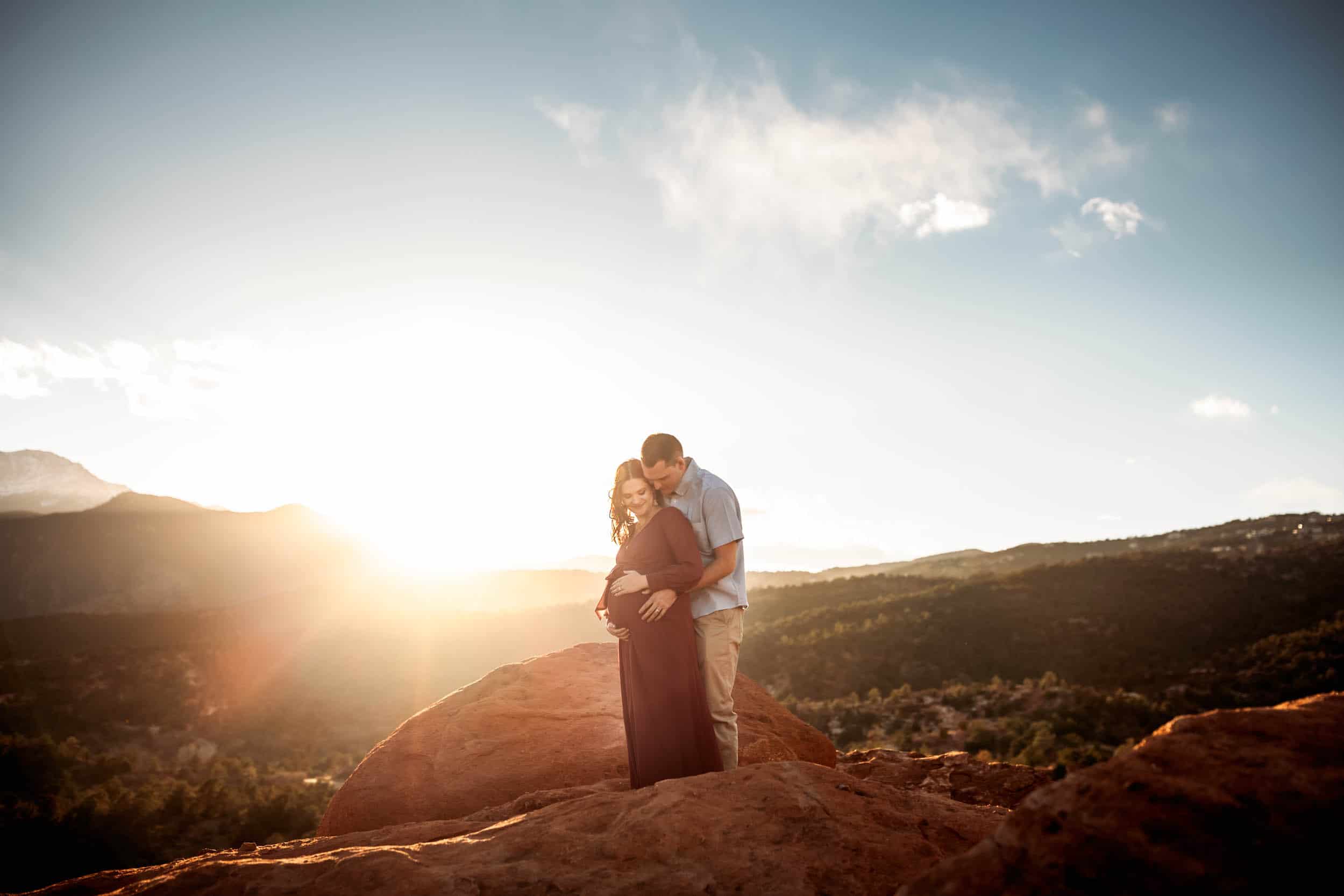 maternity couple standing on red rocks in the sunset