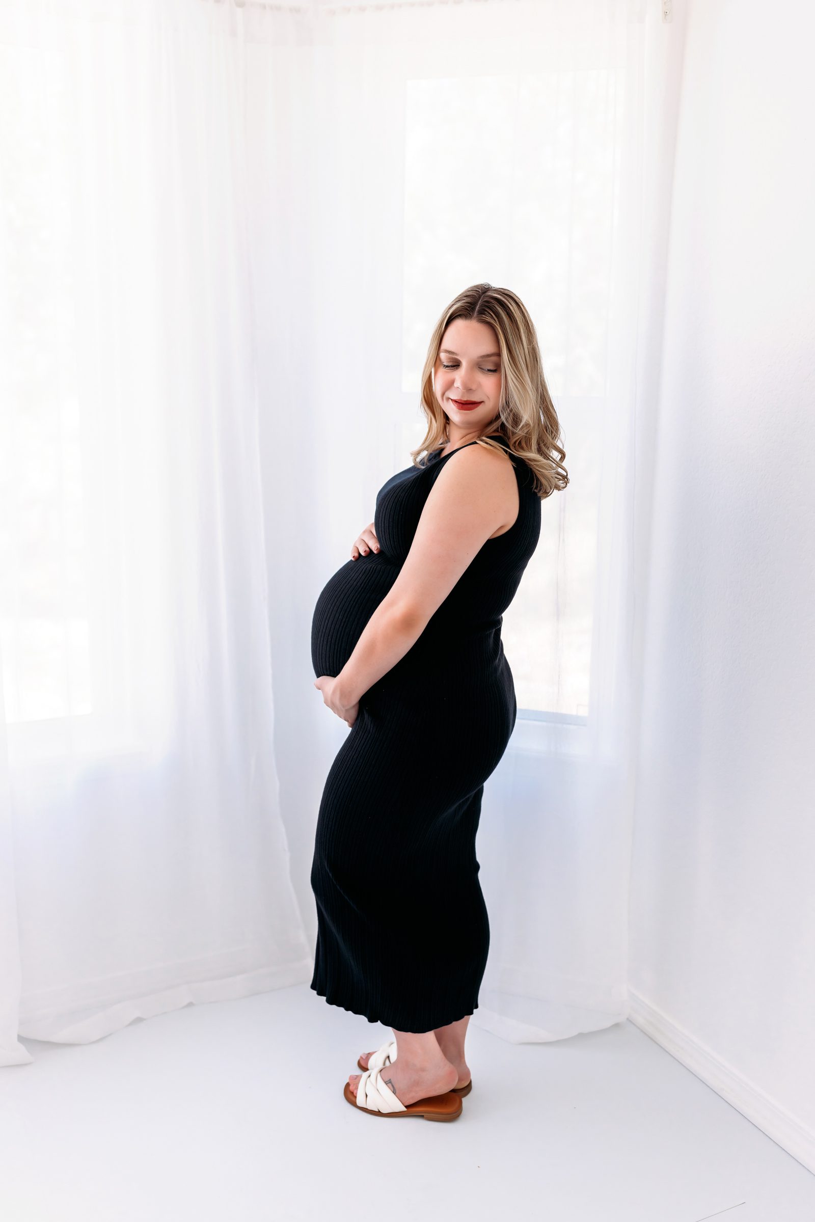 Pregnant woman in a black ribbed dress standing in a studio, gently cradling her baby bump