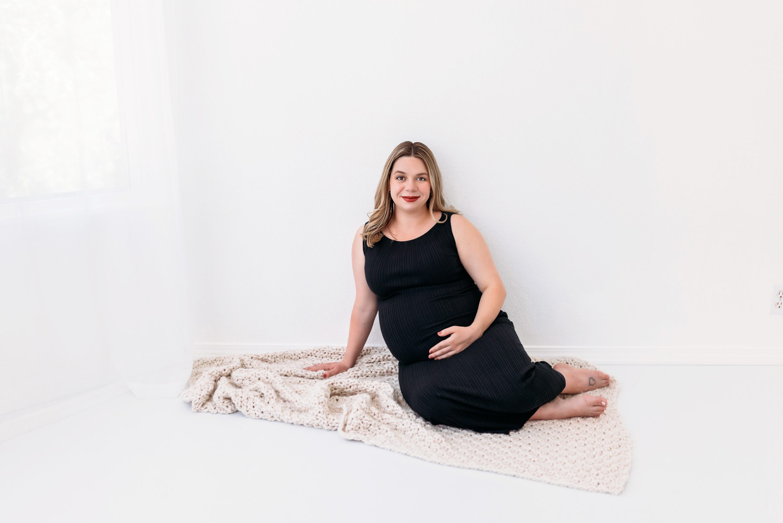 Pregnant woman in a black dress sitting on a cozy blanket, showing what to wear for maternity photos indoors