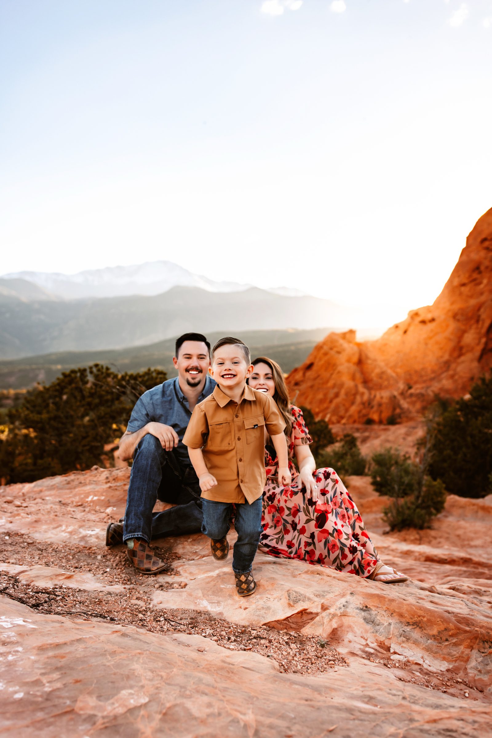 A smiling boy runs towards the camera with his parents behind him, framed by golden light in a spring photo session in Colorado Springs.