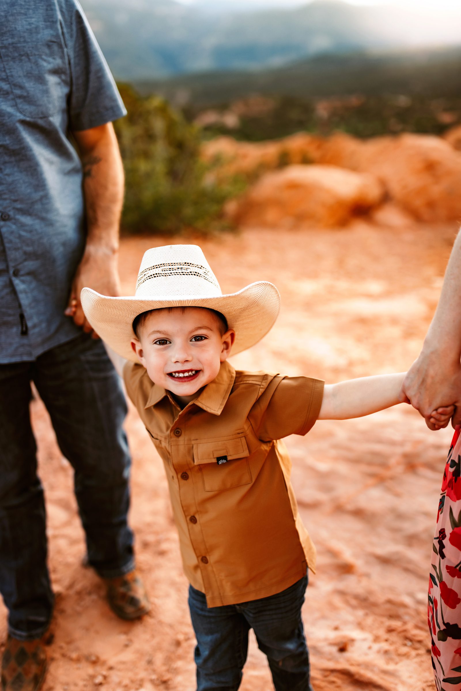 A joyful young boy in a cowboy hat excitedly holds his parents' hands, beaming during a spring photo session in Colorado Springs.