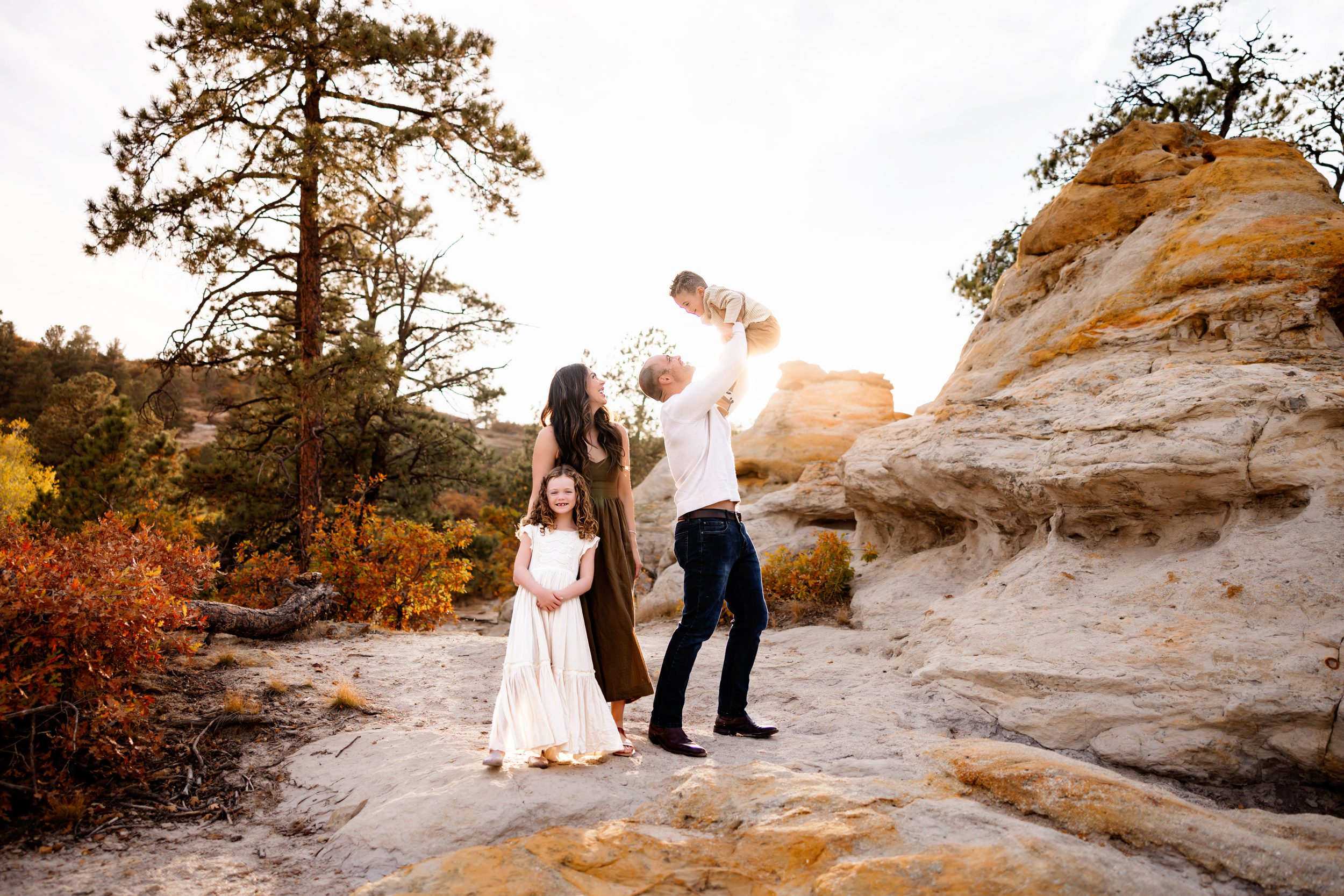 father lifts his young son into the air while the mother and daughter look on with smiles, framed by breathtaking natural rock formations and fall colors.