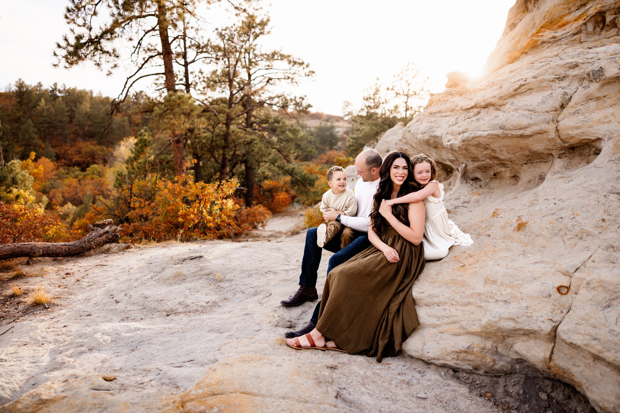 A family sits nestled against a sandstone rock formation, sharing laughter and warm embraces in the golden autumn light