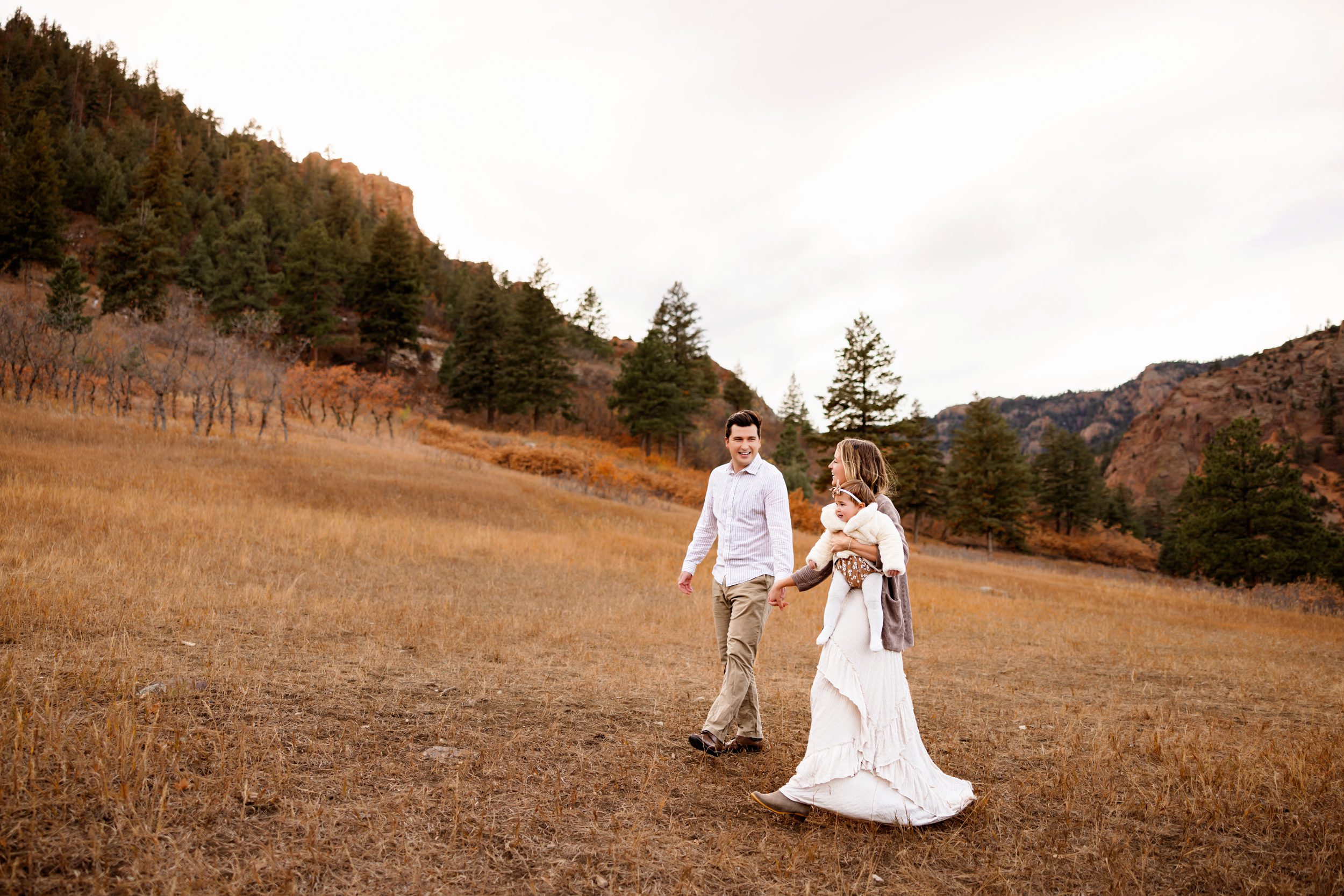 The family walks through a golden field, holding hands and soaking in the beauty of the Colorado Springs Outdoor Photography Session, framed by stunning mountain views