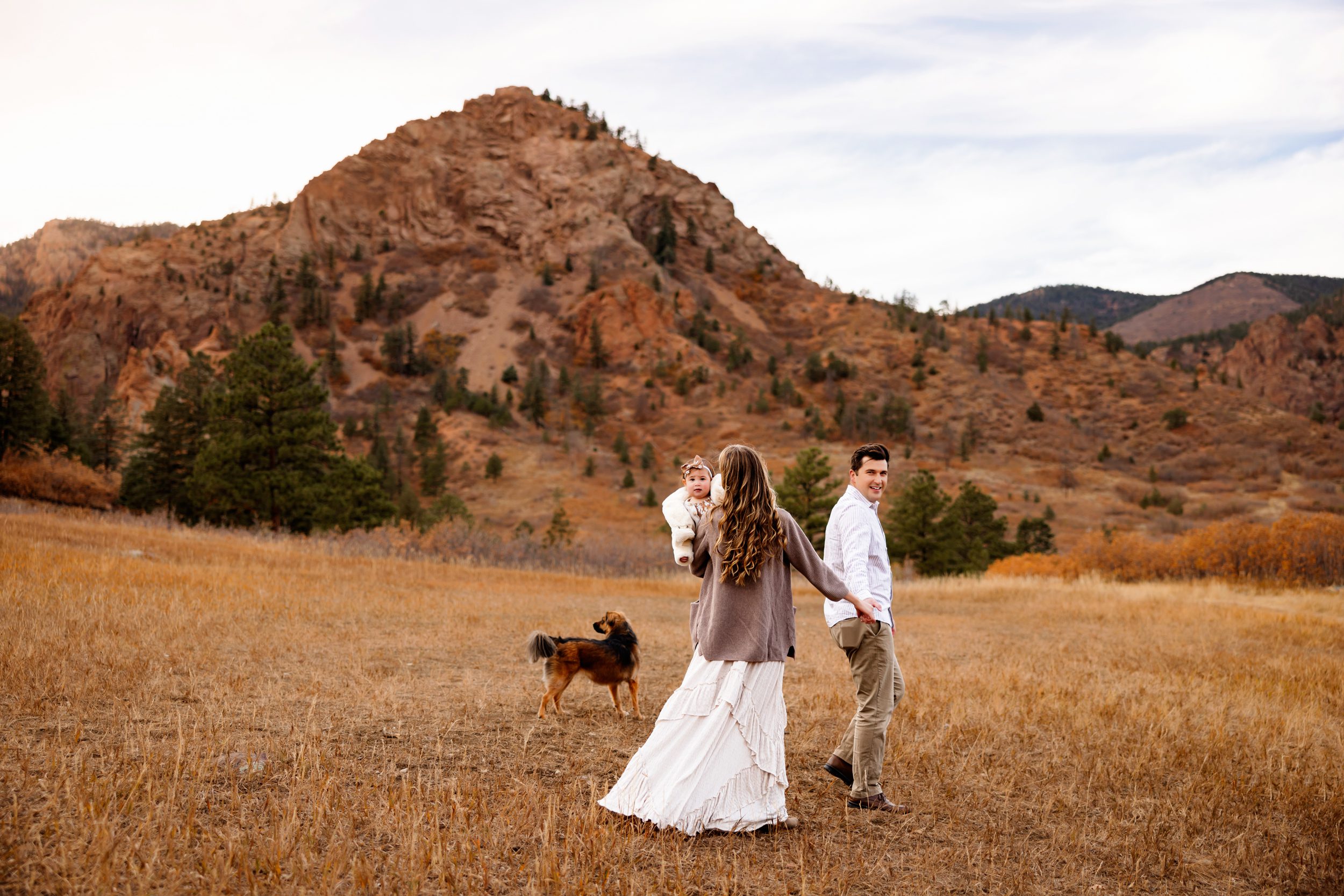 A couple strolls hand in hand through an open field, their baby snuggled in her mother’s arms, creating a timeless moment in their Colorado Springs Outdoor Photography Session