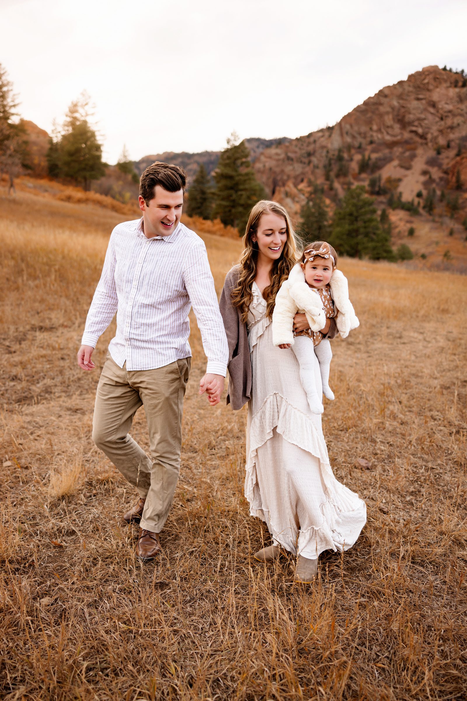 A couple beams with happiness while holding their baby during a Colorado Springs Outdoor Photography Session, framed by rolling hills and vibrant fall colors.