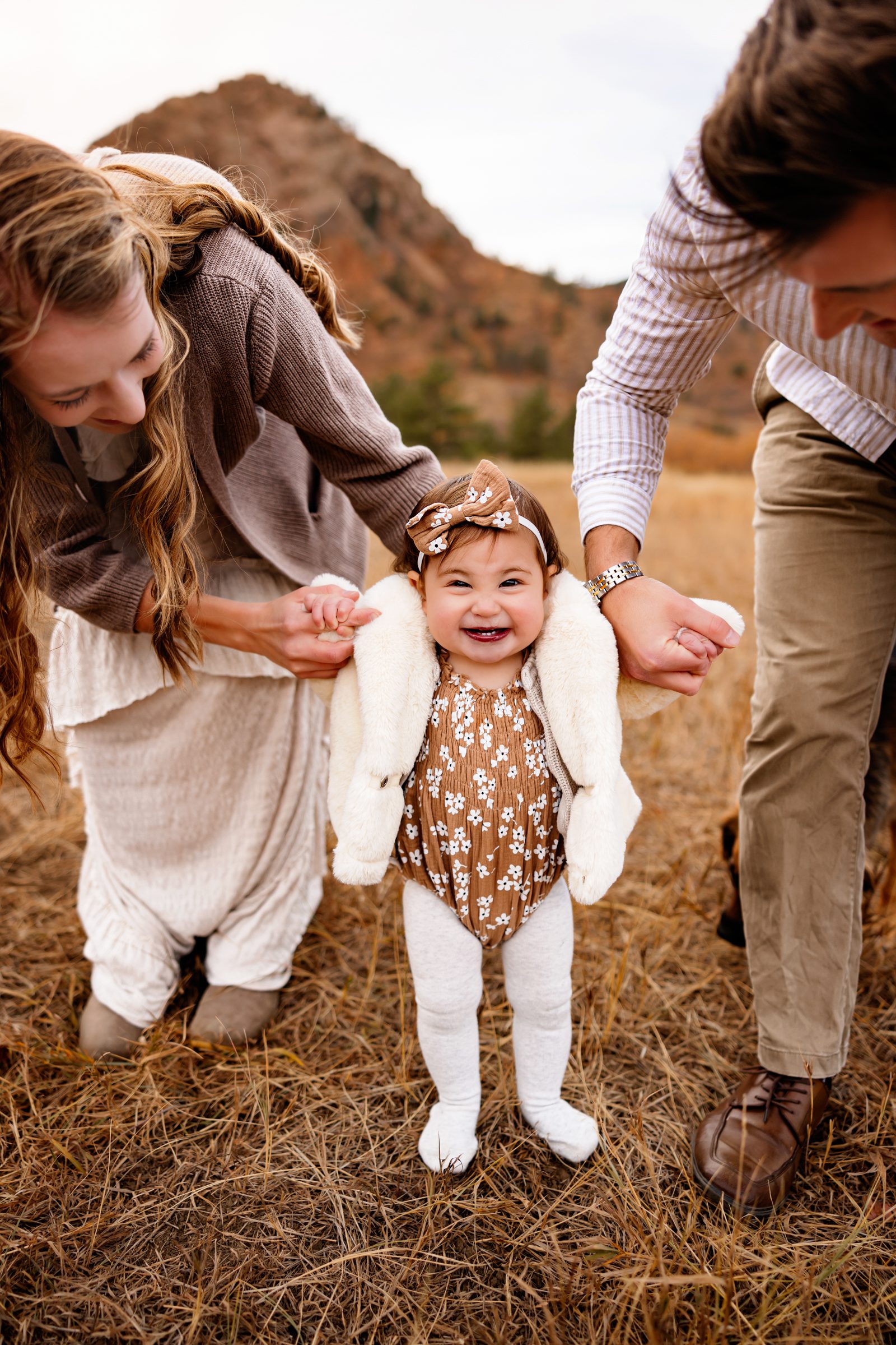 Parents hold hands with their baby girl while she gives a big smile