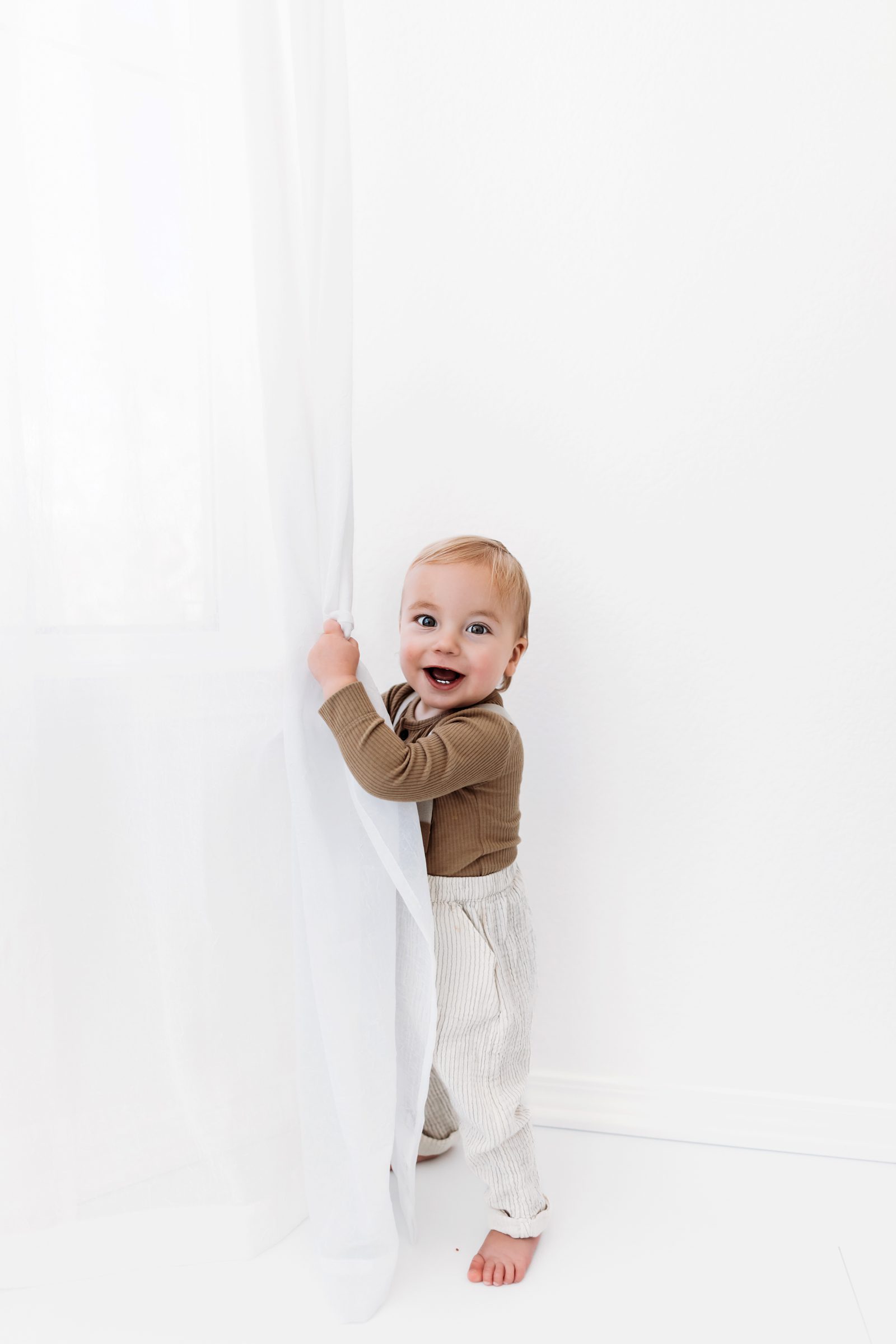Baby standing near a white curtain, smiling playfully.