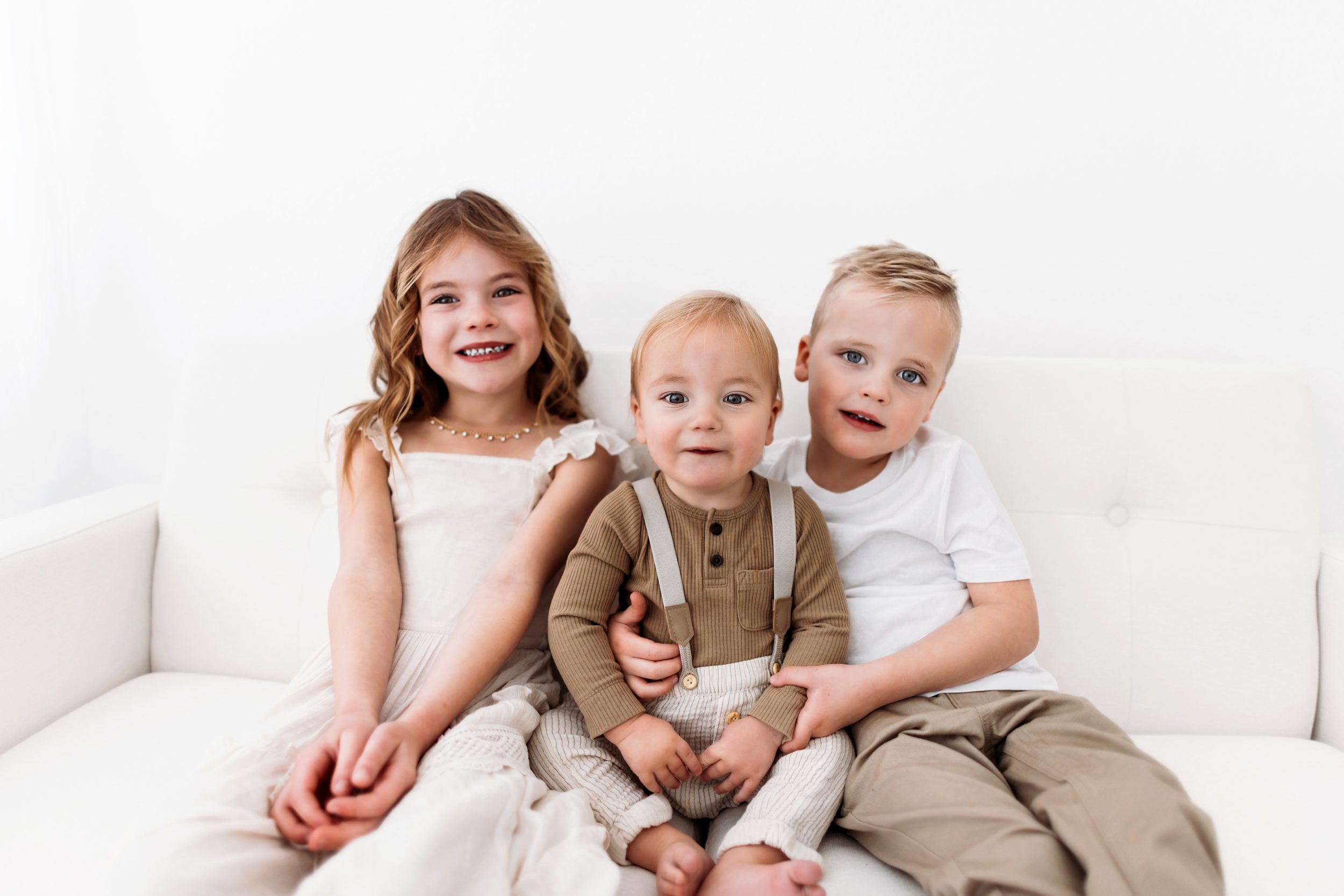 Three siblings sitting close on a white couch, smiling at the camera