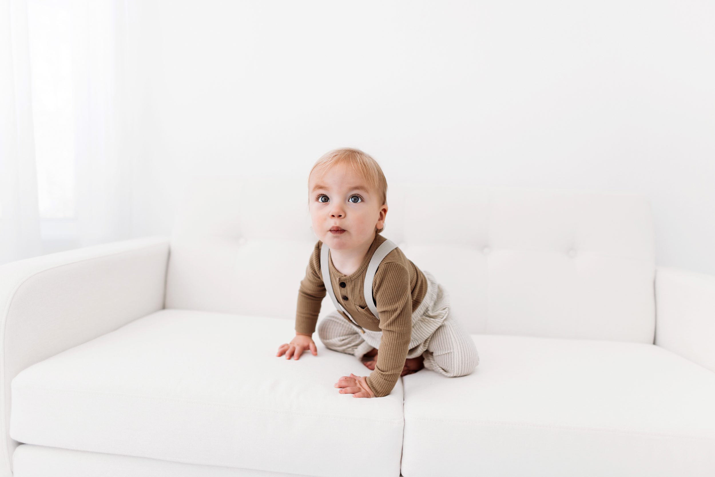 Toddler sitting alone on a white couch in neutral-toned outfit at Sibling studio session in Colorado Springs