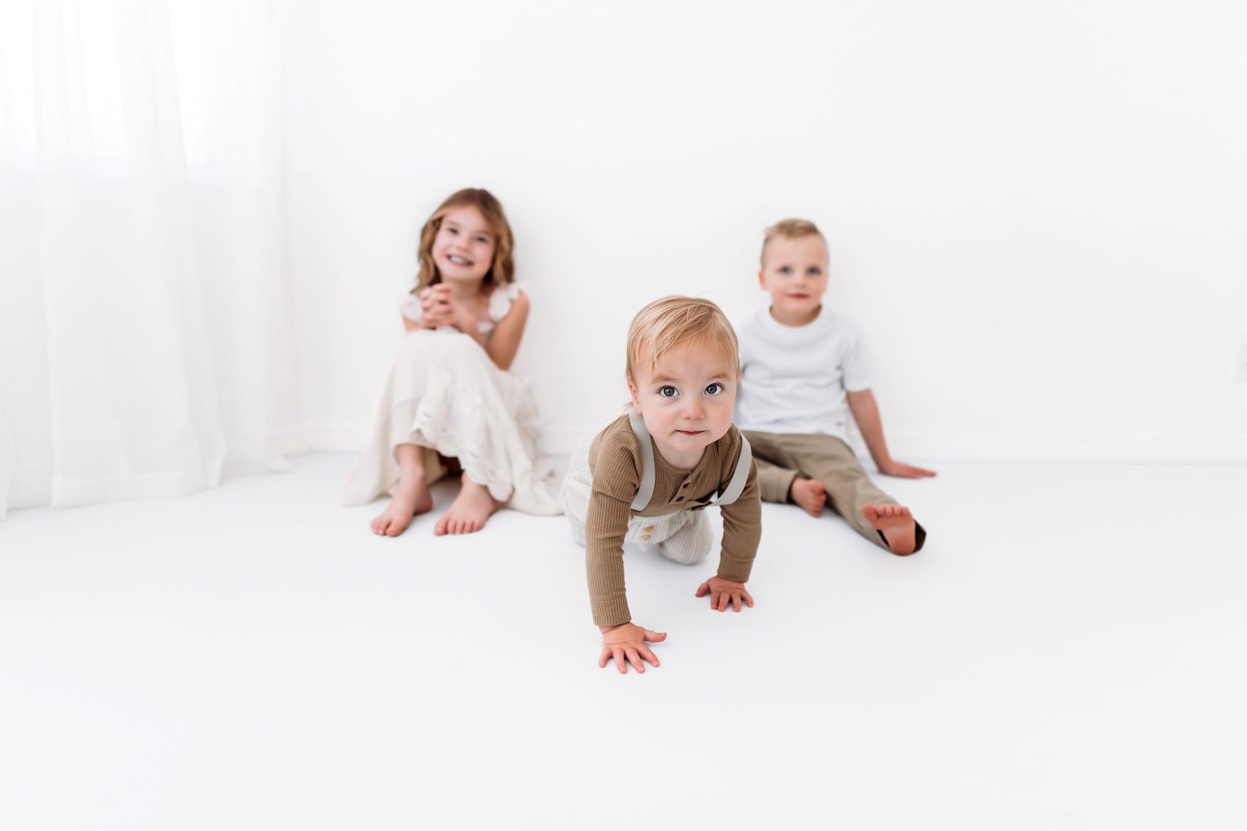 Baby crawling toward the camera with siblings smiling in the background.