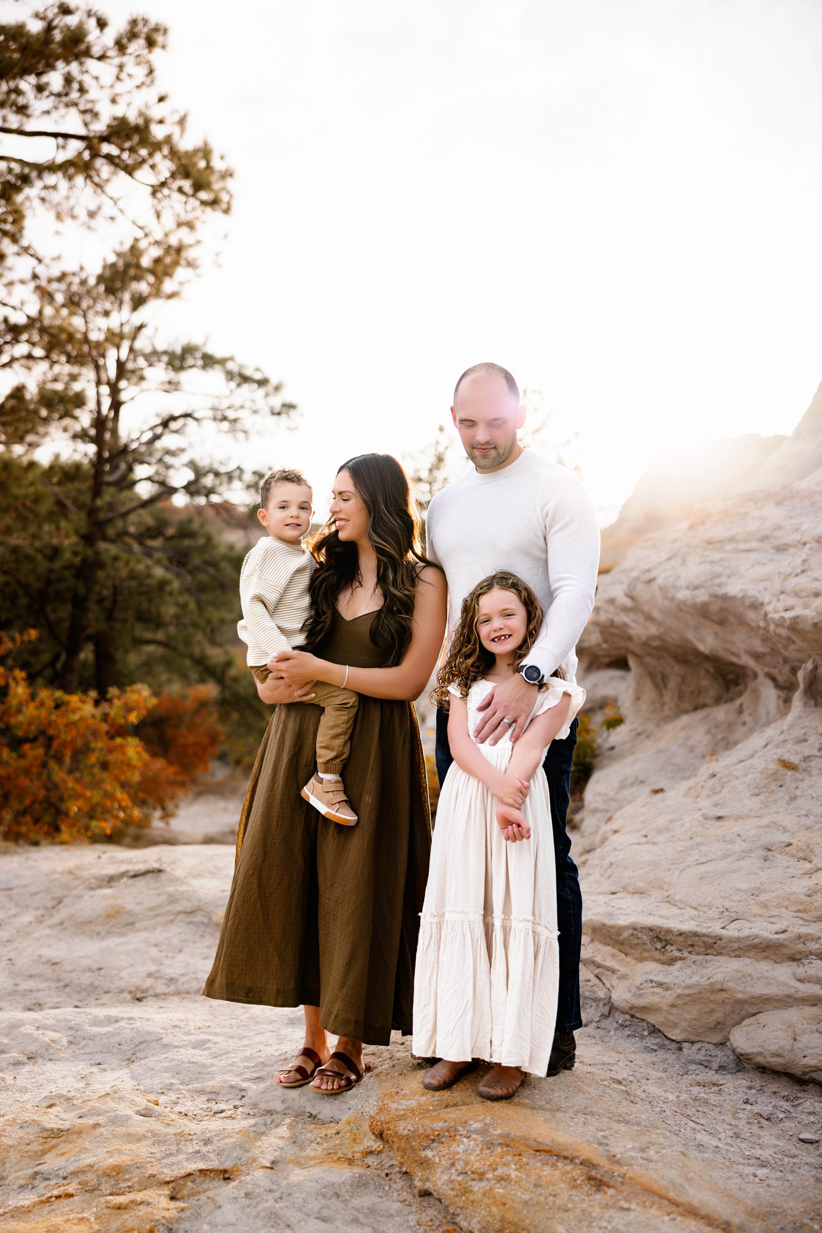 A family of four poses together on a rocky landscape with golden sunlight behind them, the mother holding their young son while the father stands beside their daughter