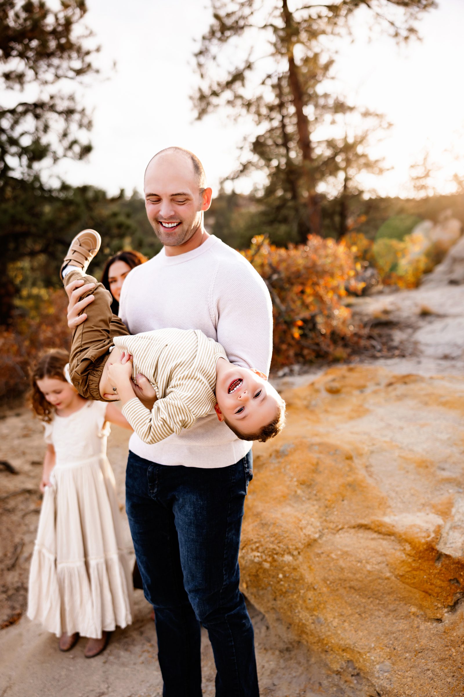A father laughs while holding his young son upside down, as autumn leaves and warm evening light create a beautiful outdoor setting.