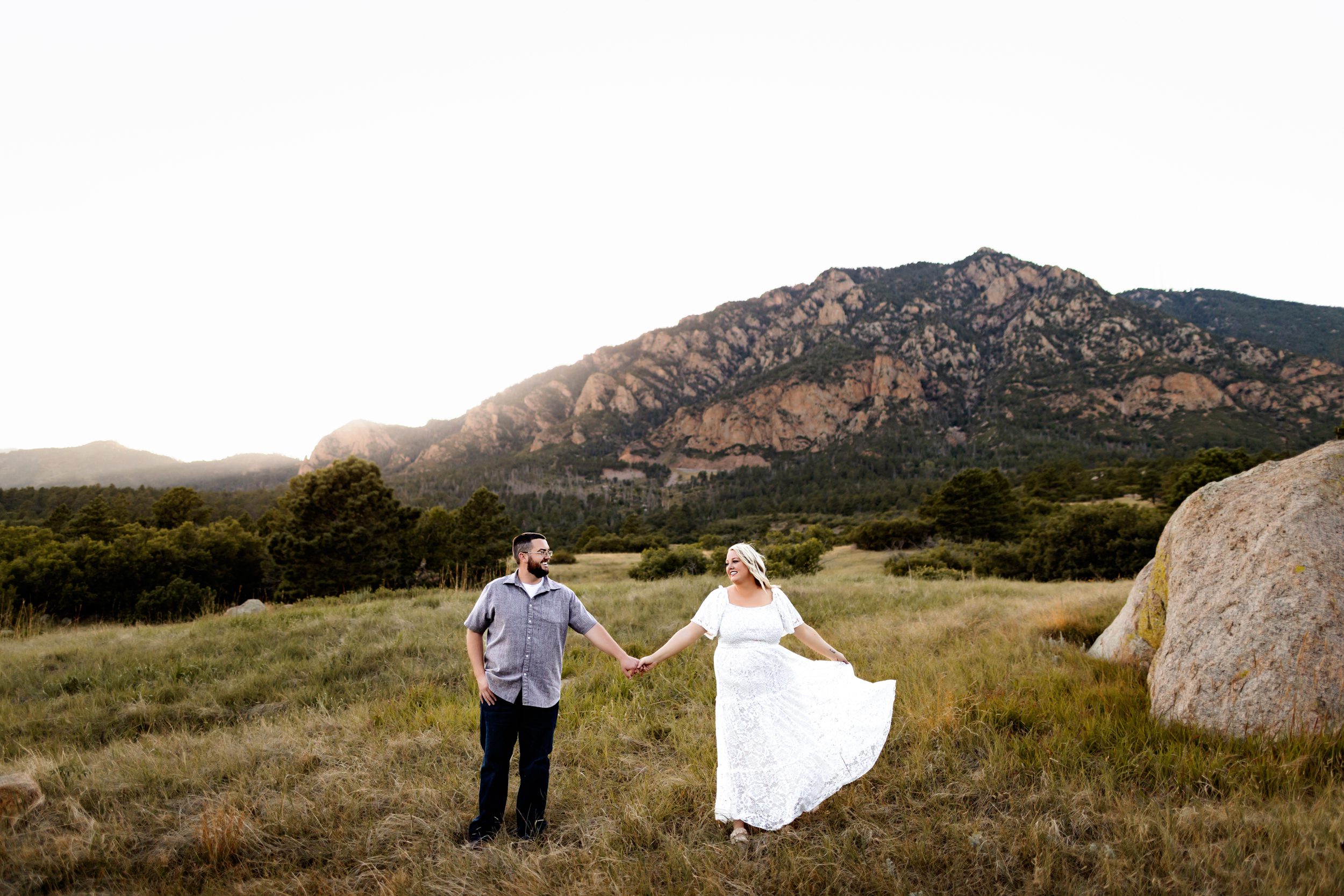 A joyful couple walking hand-in-hand through an expansive field, with stunning Colorado Springs mountains towering behind them during their outdoor session.