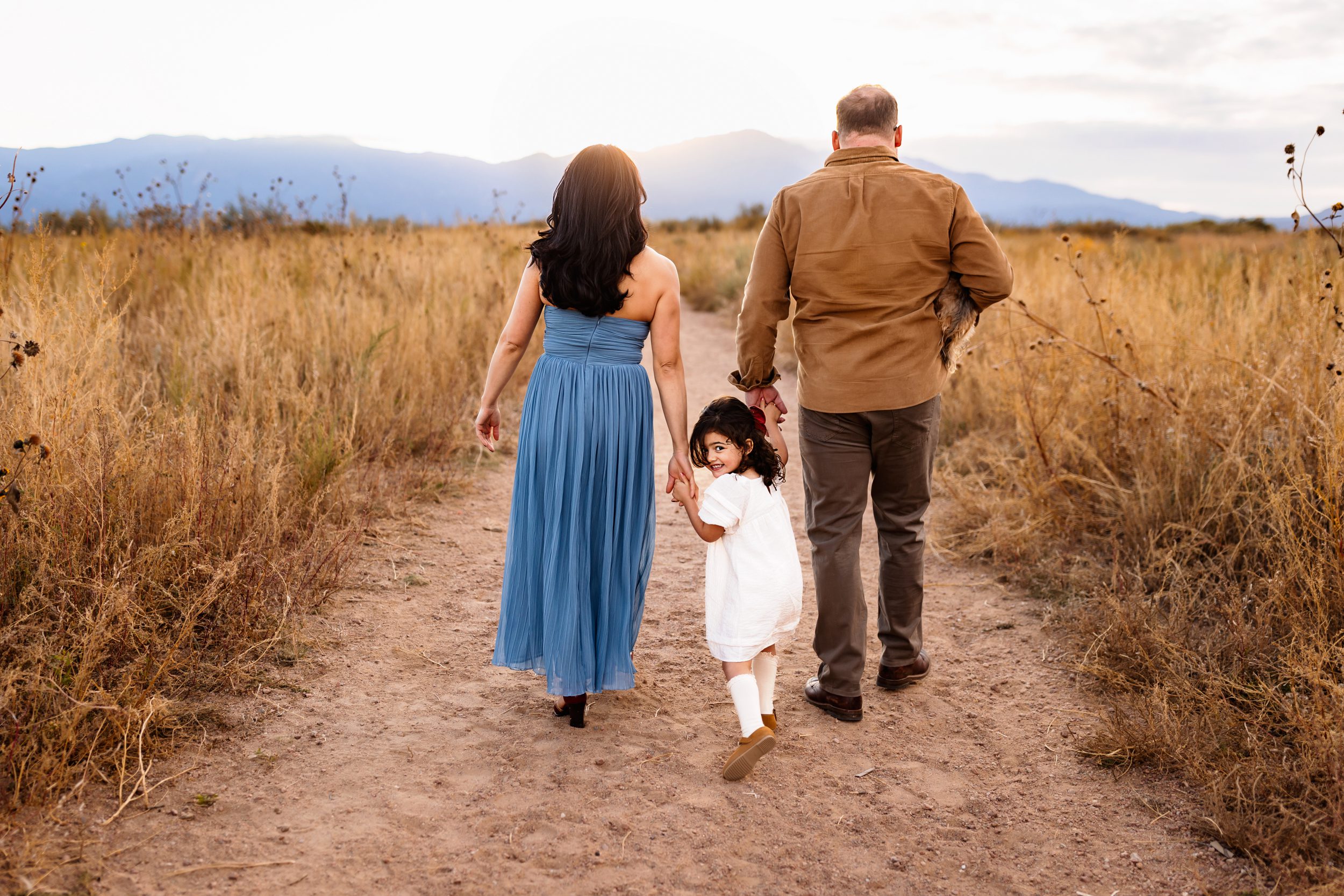 A family of three walks hand in hand while their little girl glances back with a sweet smile