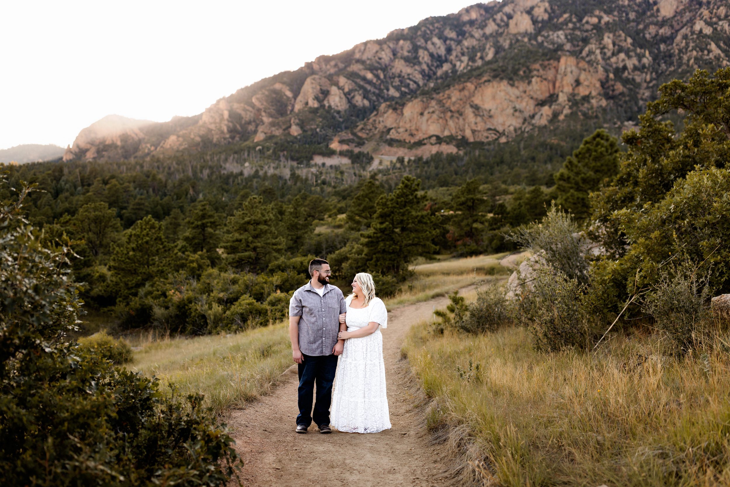A couple walking hand-in-hand down a dirt path surrounded by pine trees and dramatic mountain views at sunset