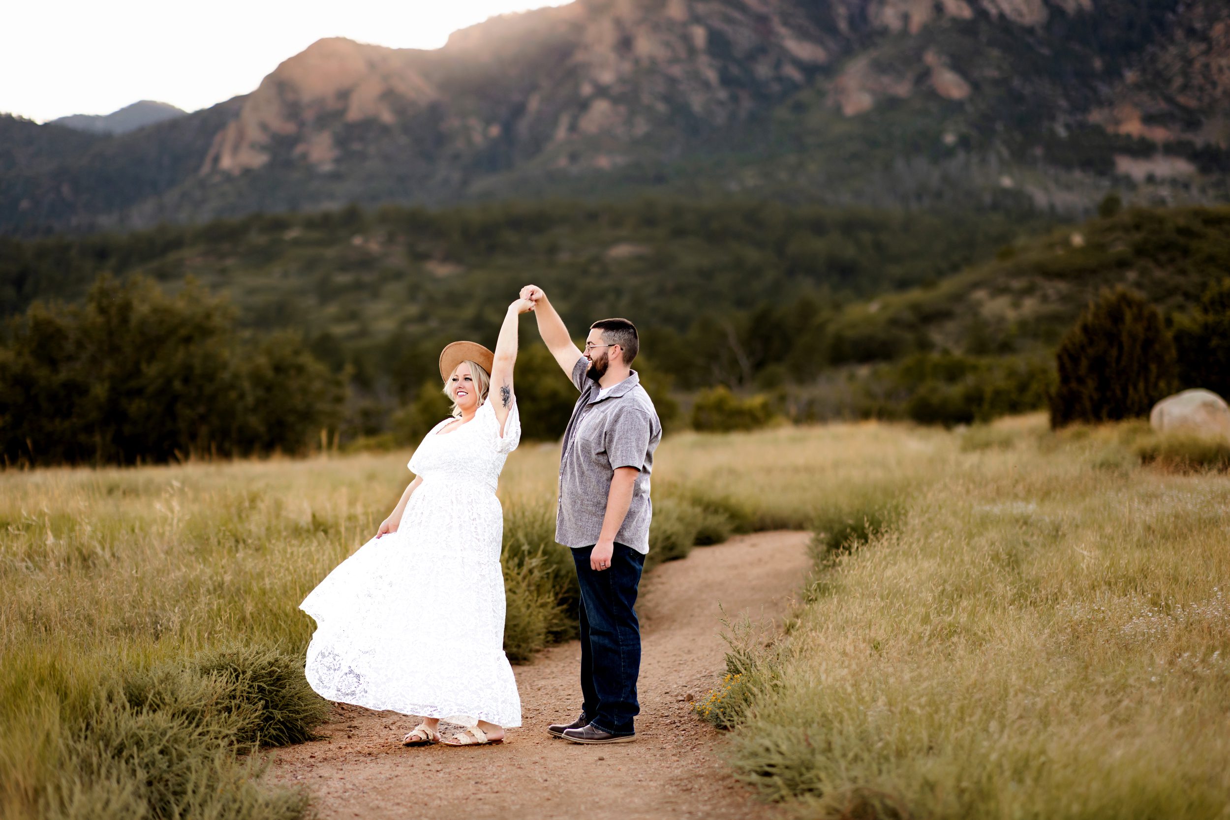 A playful twirl as the couple dances on a dirt path surrounded by tall grass and sunset-lit peaks in the background