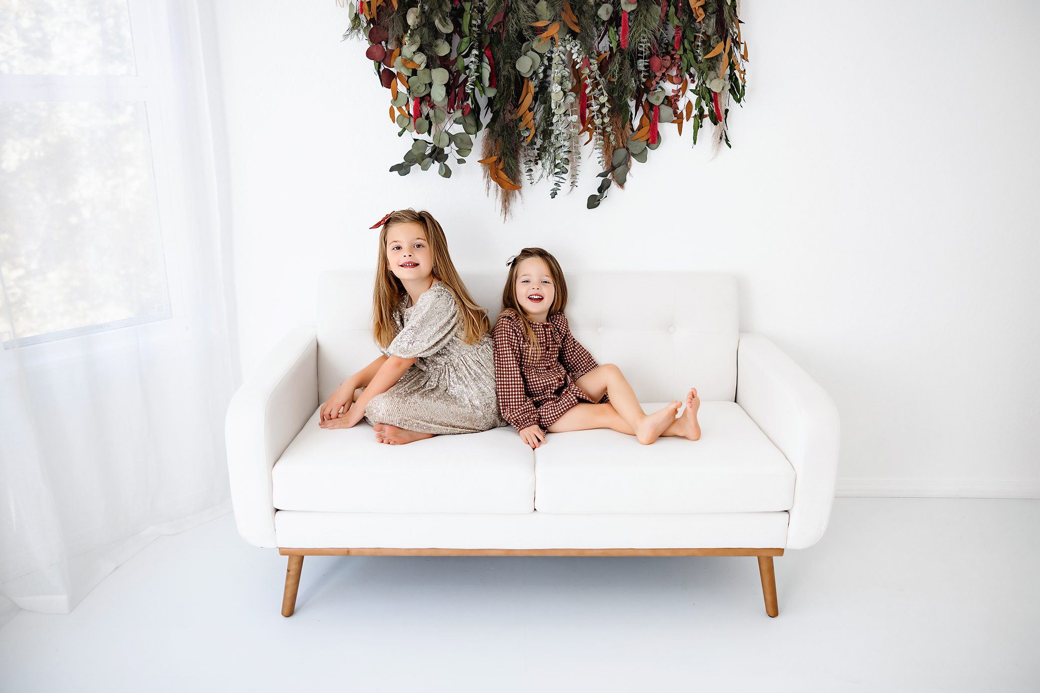 Two young girls in festive dresses smiling and sitting back-to-back on a white couch with a holiday greenery backdrop