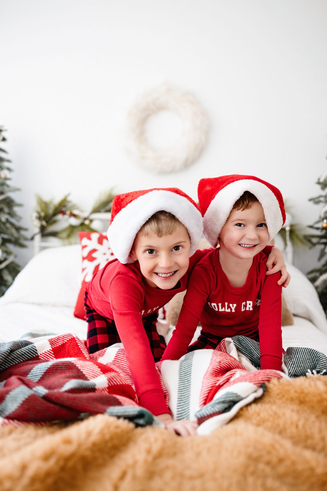 Two boys in red pajamas and Santa hats smiling on a cozy holiday bed with plaid blankets at a Colorado Springs Indoor Holiday Mini Session