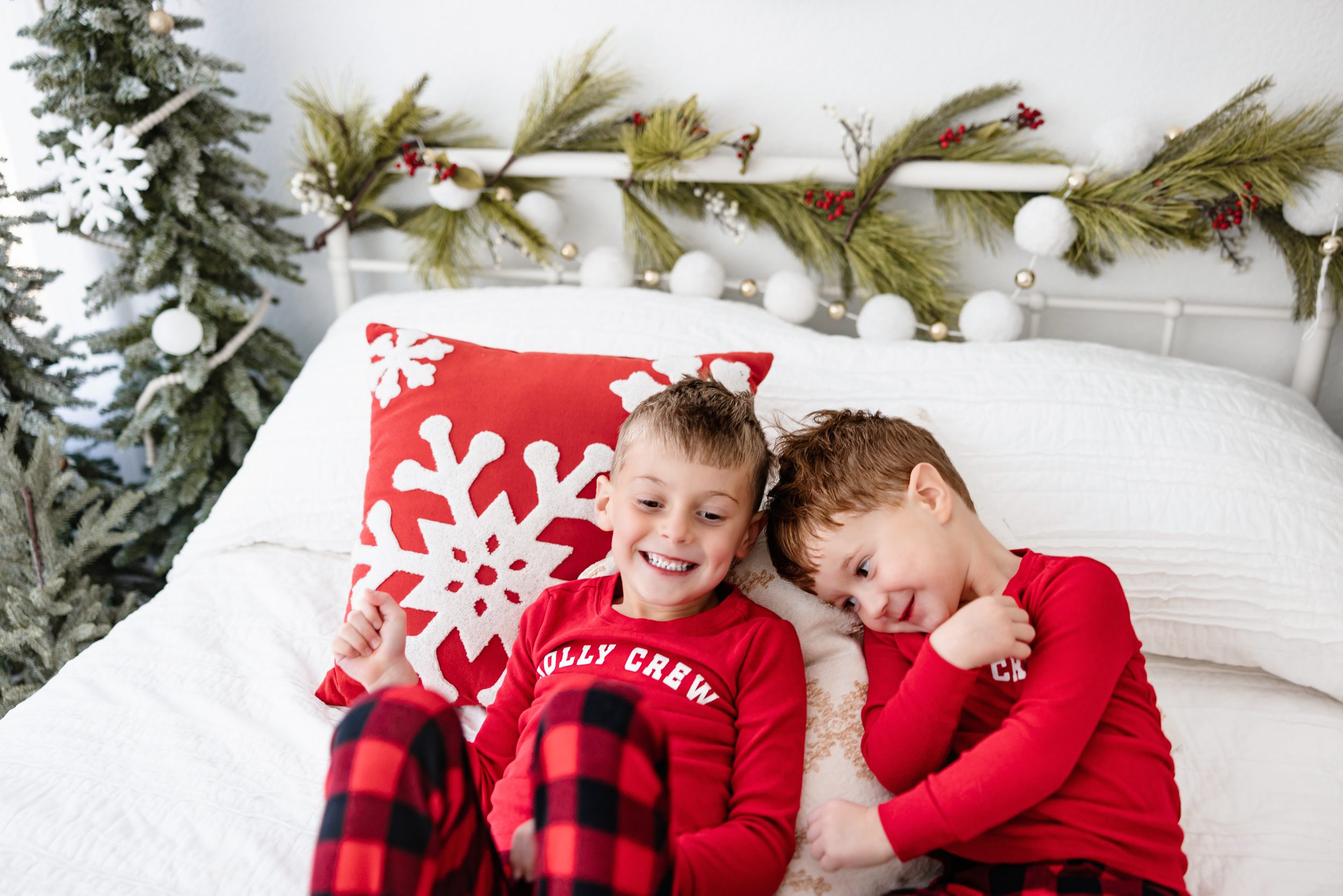 Two young boys in matching red holiday pajamas laughing on a festive white bed with snowflake pillows and evergreen garland