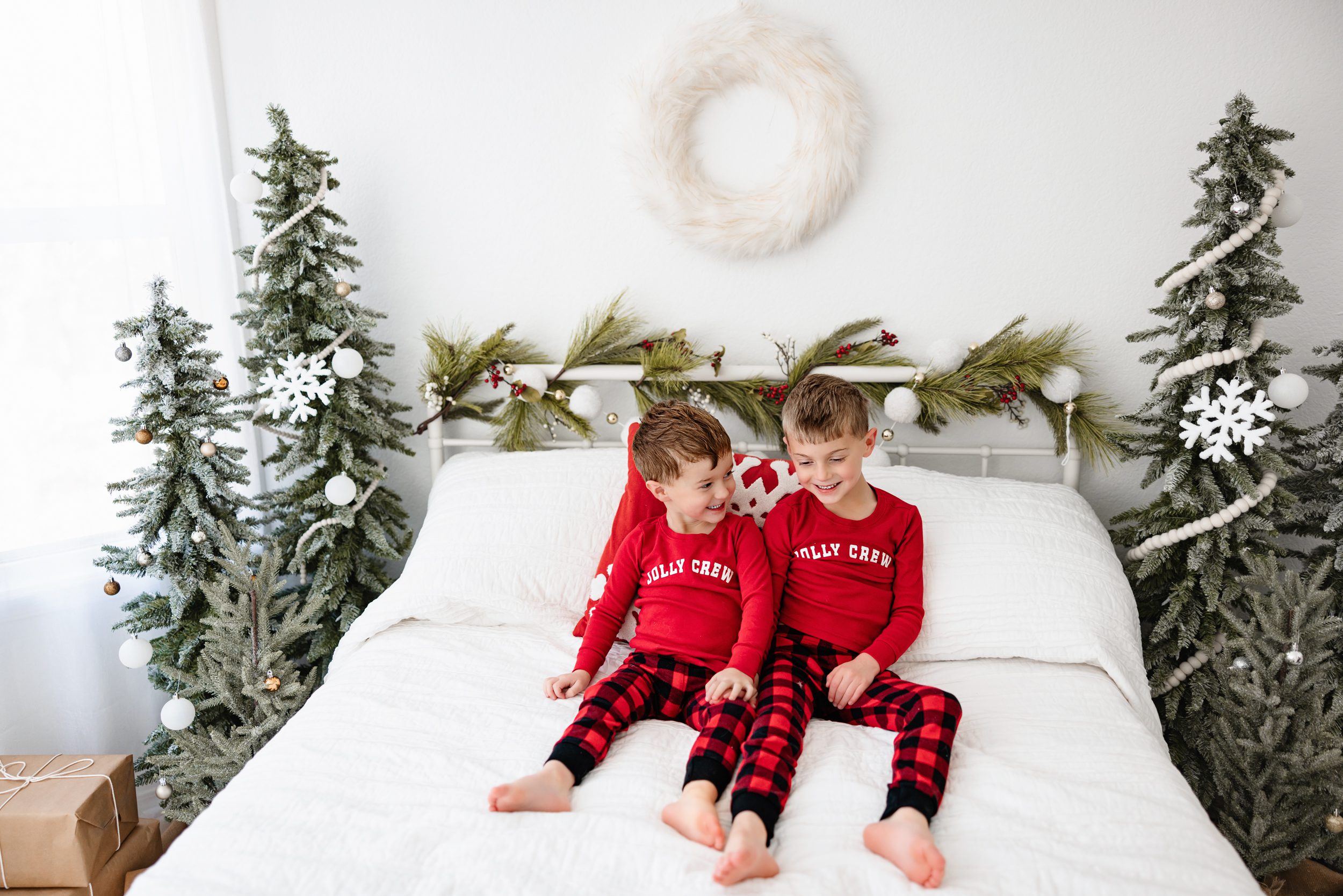 Two boys in red “Jolly Crew” pajamas sitting on a holiday-decorated white bed, surrounded by frosted Christmas trees and wrapped gifts