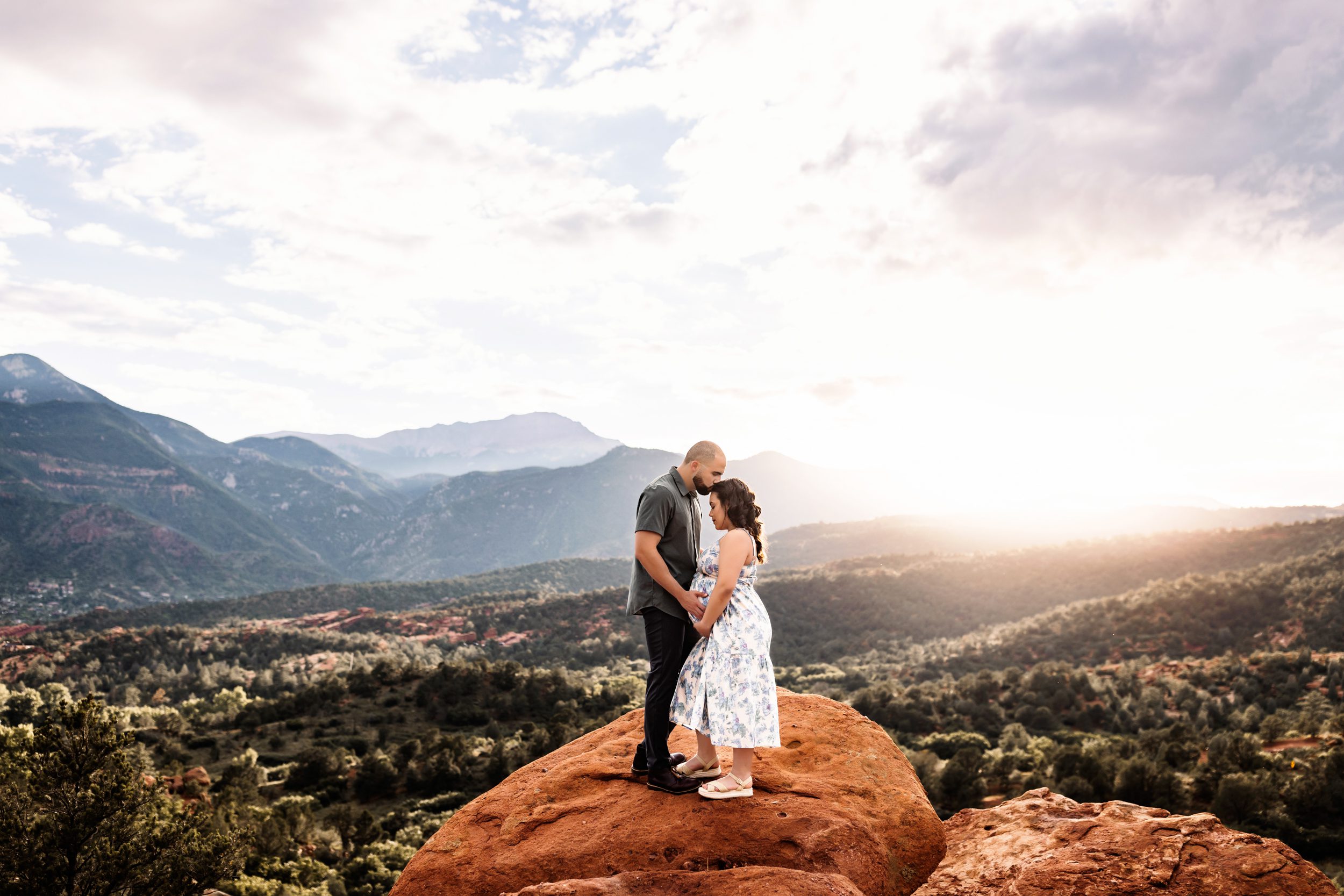 Couple embracing on a red rock ledge during a Colorado Springs outdoor maternity session with mountains in the background