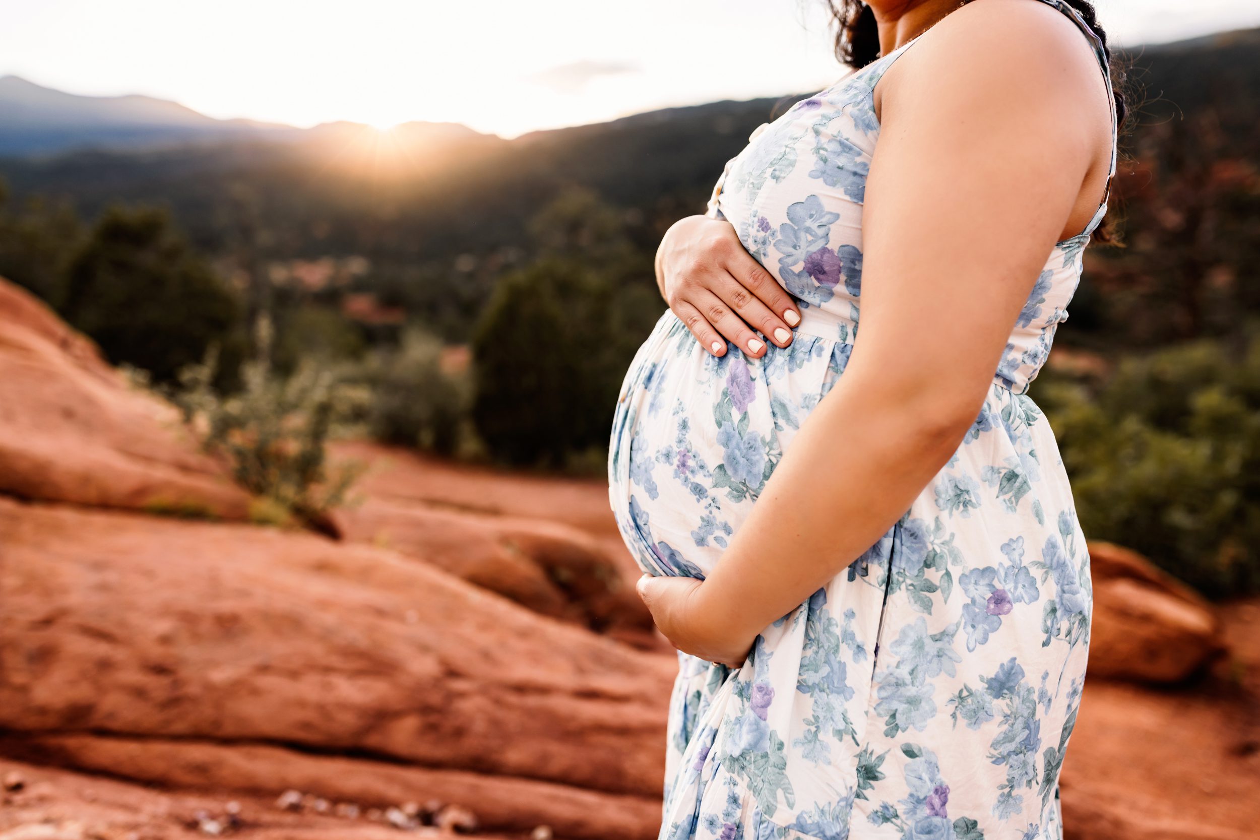 Detail shot of a pregnant belly framed by hands, with a blurred sunset and red rock background