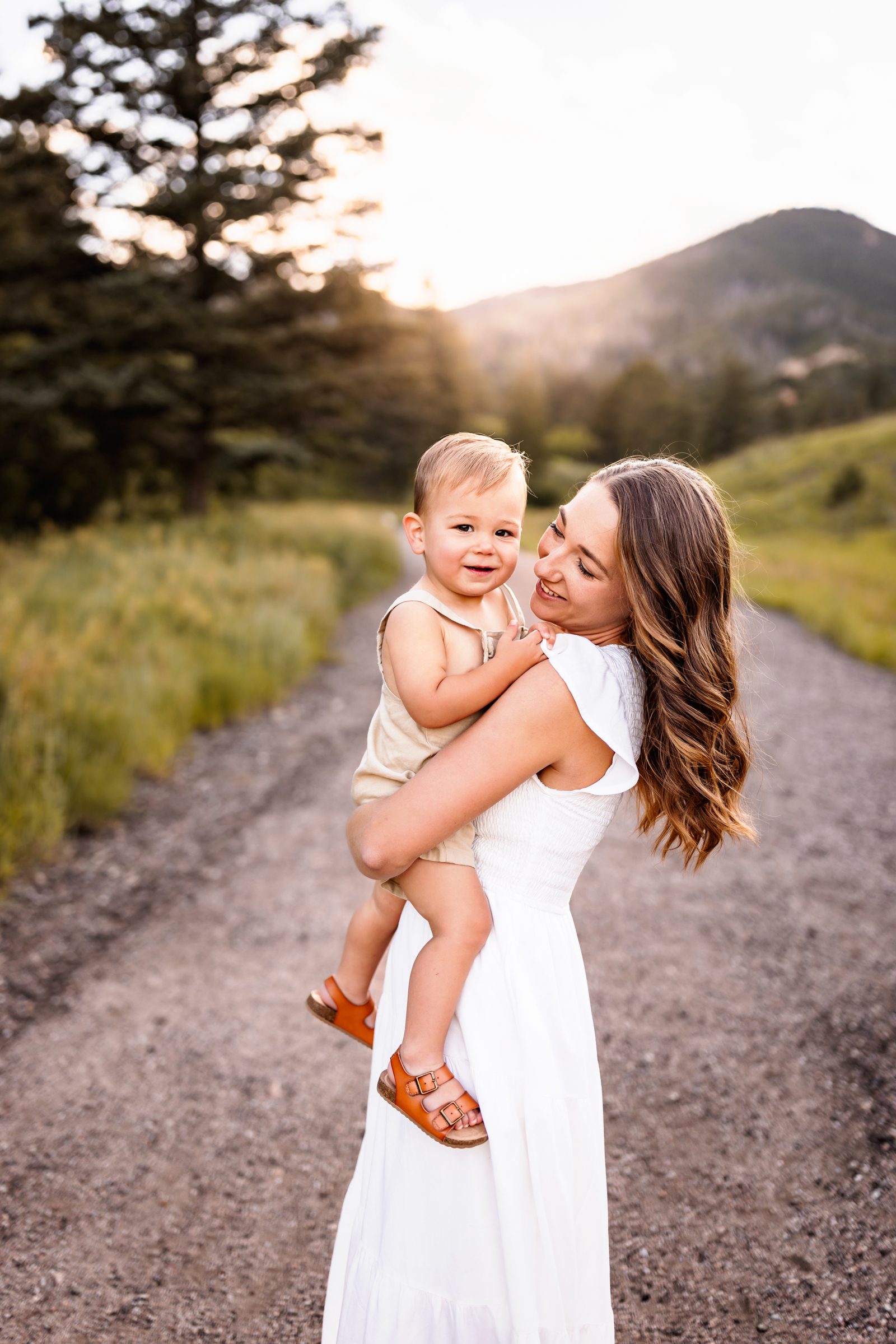A mom and a young toddler boy dressed in neutral colors with the setting sun and mountain behind them