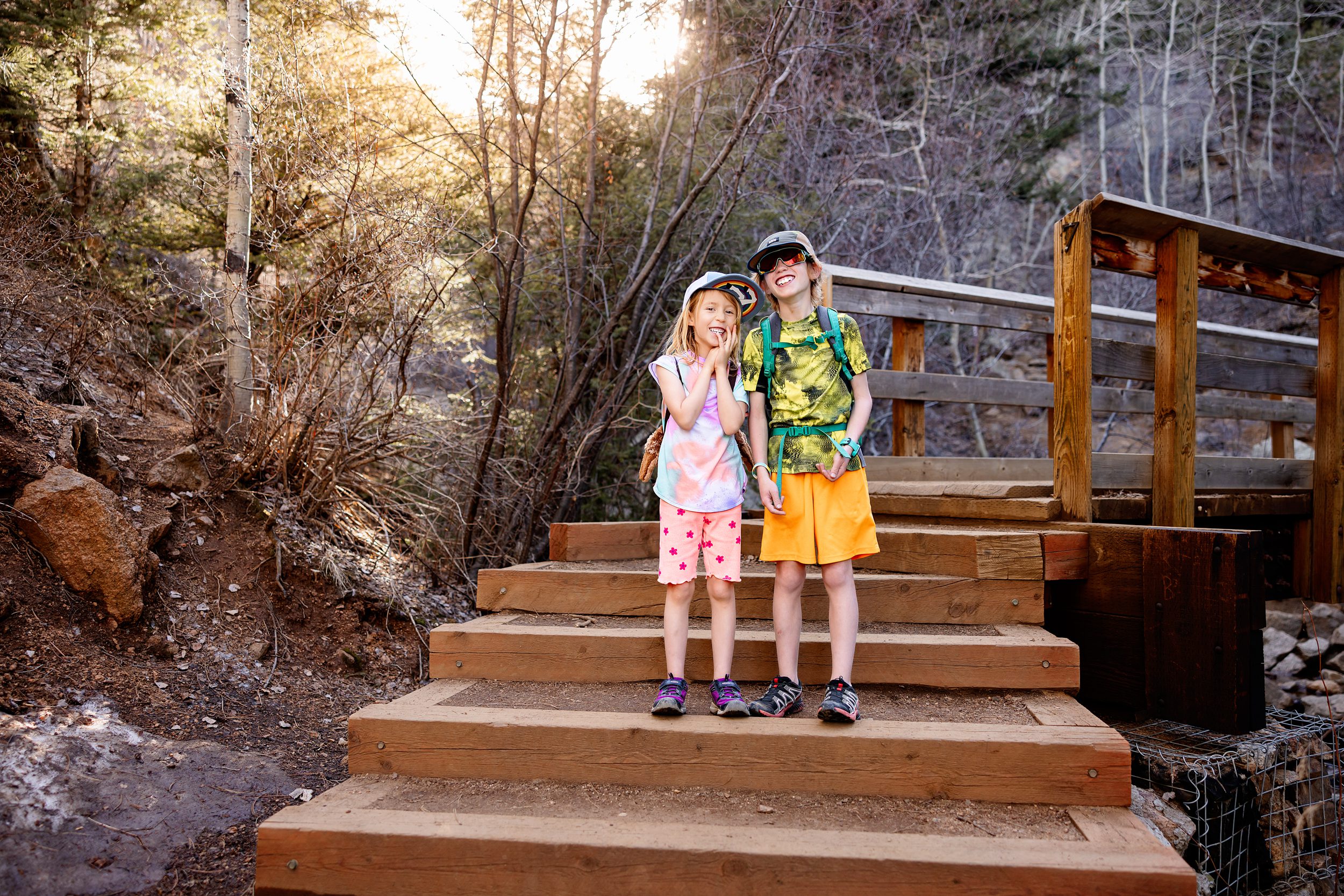 Brother and sister stopping for a photo break on a wooded hiking trail with evening sun behind them.