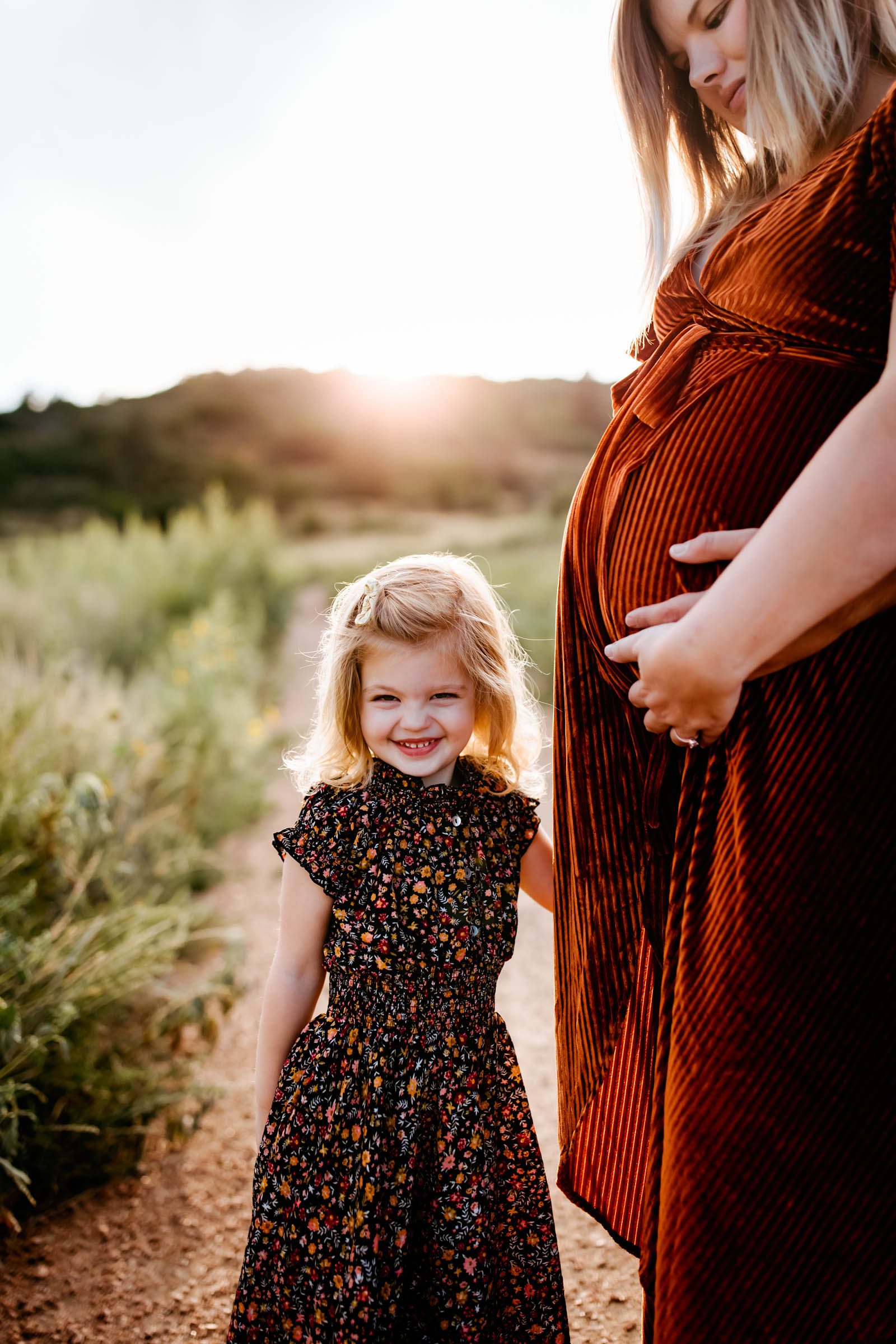 Pregnant moms belly and toddler girl smiling at the camera with the setting sun behind them