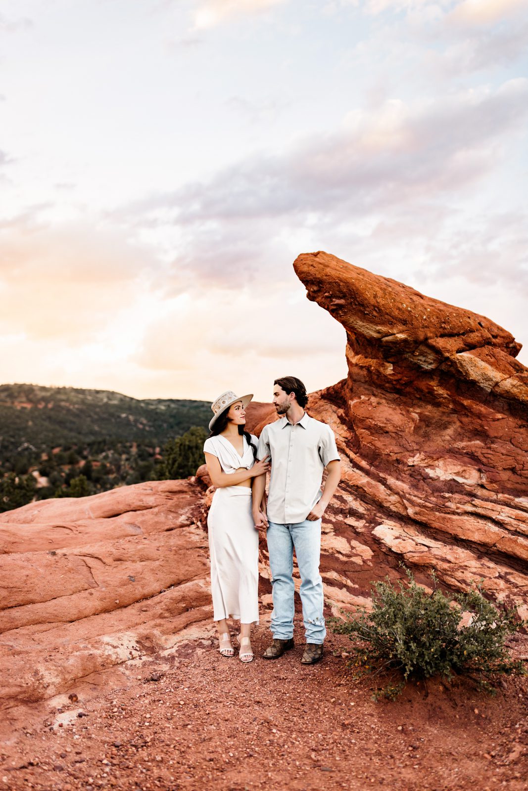 Stylish couple standing near a large red rock formation as the sky glows with soft sunset colors.