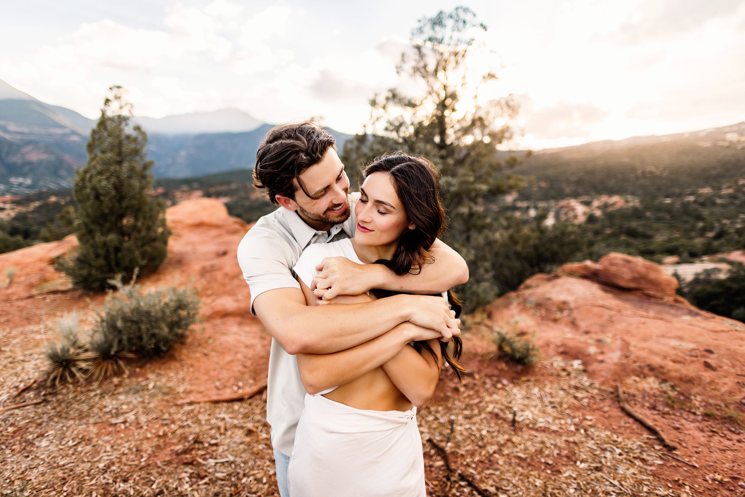 Couple wrapped in a cozy embrace with soft sunset light filtering through the trees and rock formations.