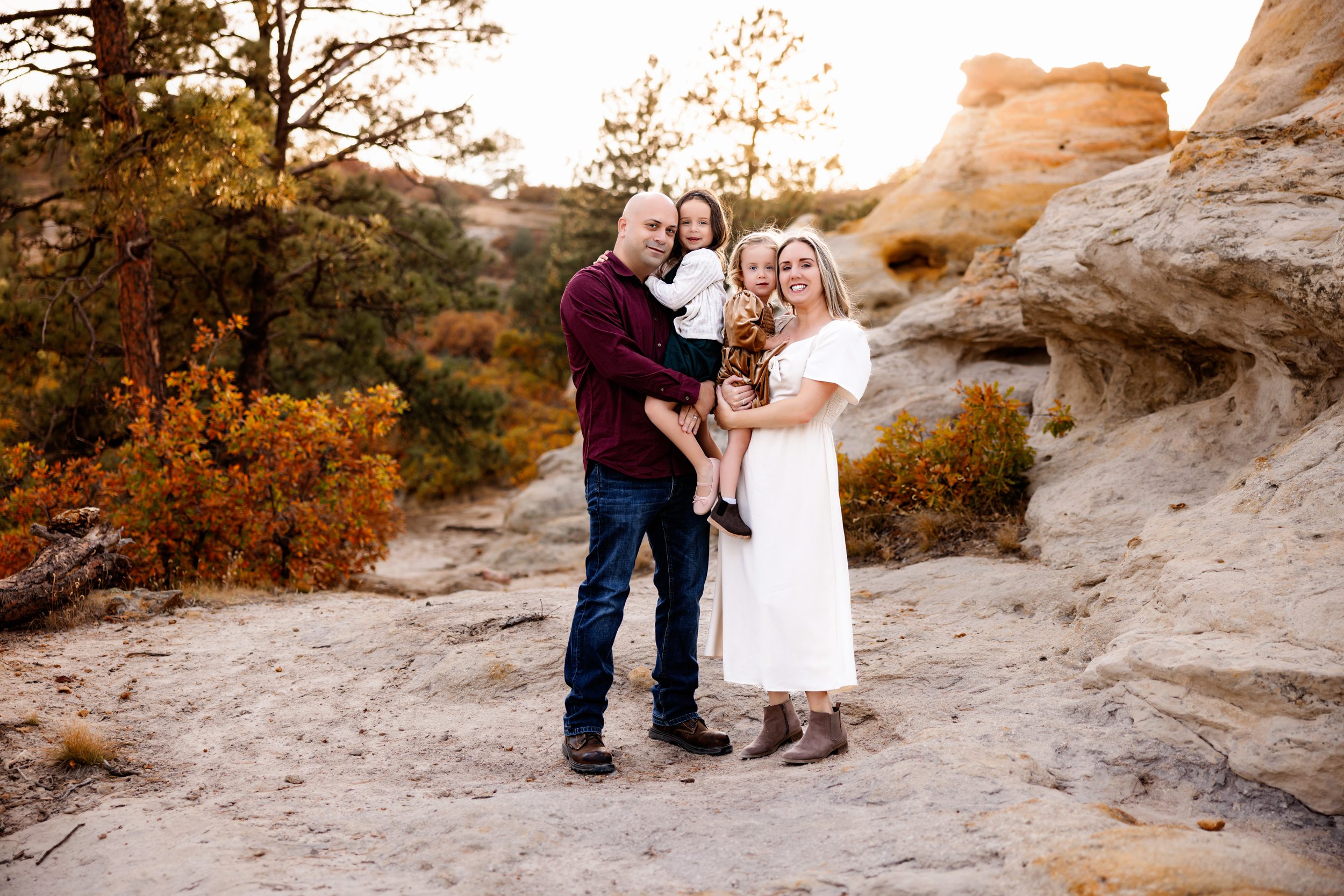 A family of four smiles together during a Fall Family Photo Session in a scenic Colorado location with white rock formations and autumn foliage.