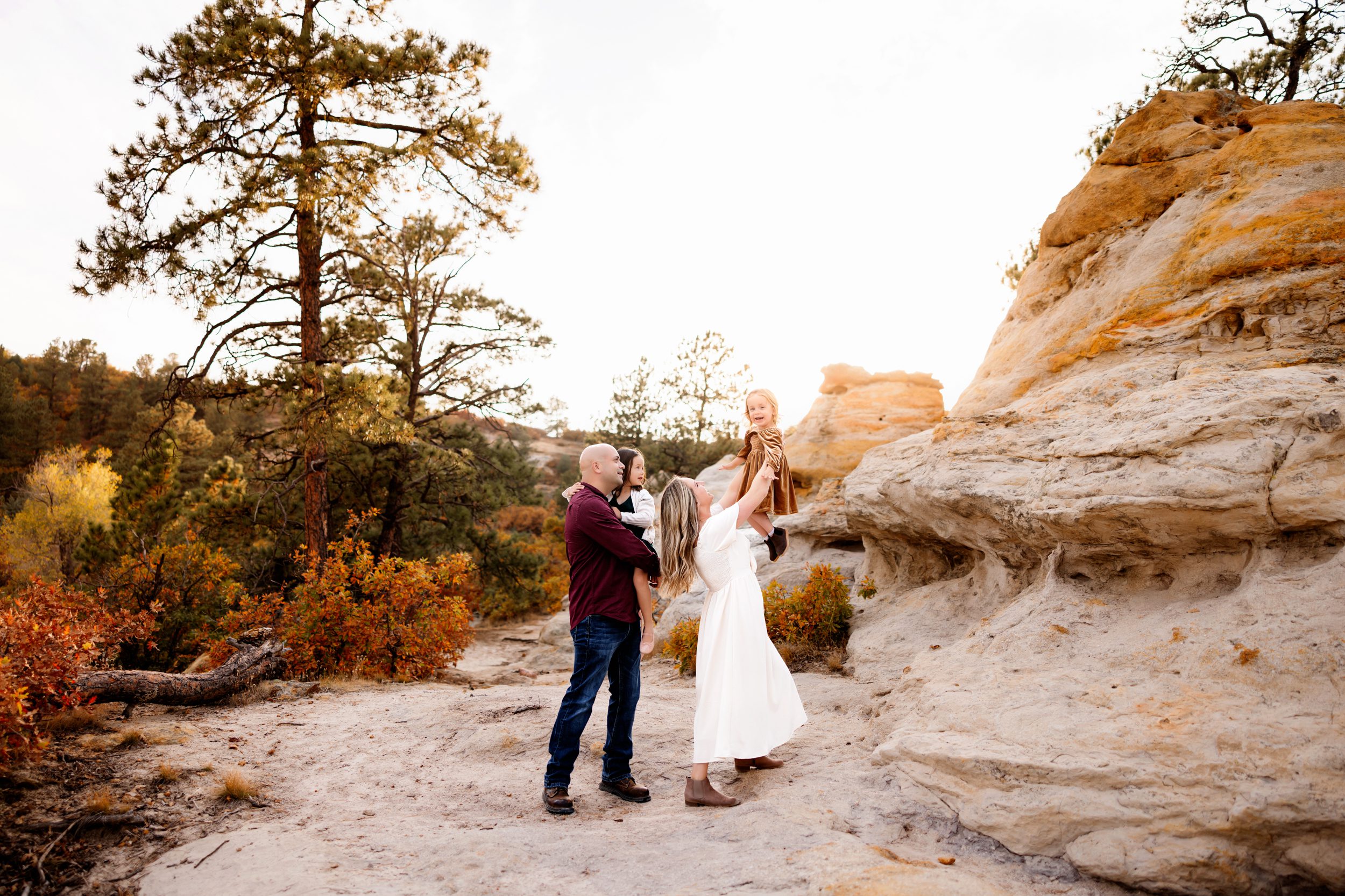 A mother lifts her toddler into the air as the family laughs together in a golden sunset among sandstone formations.