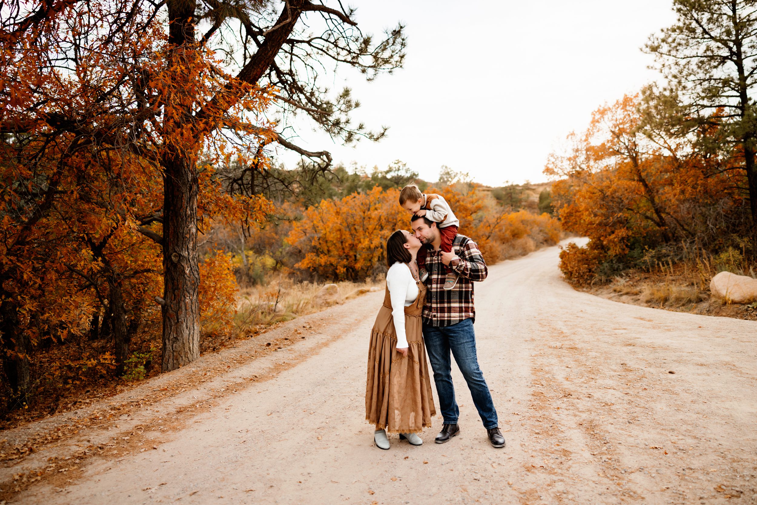 Dad holding child on shoulders while mom kisses him, framed by golden trees and a winding dirt road.