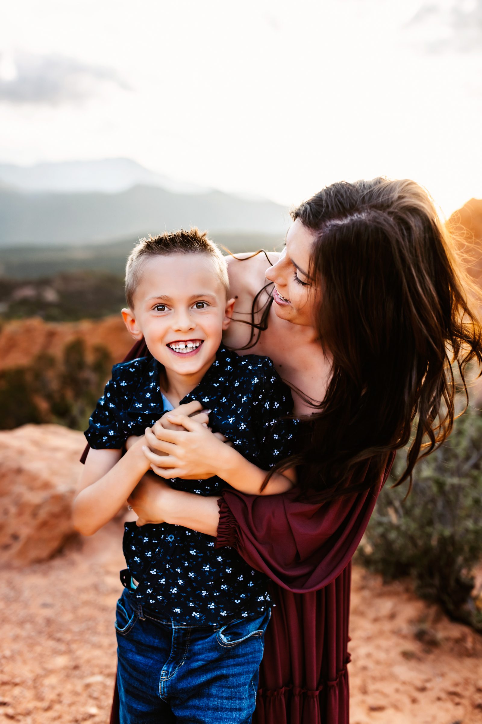 Mother hugs her son tightly as they smile with golden sunlight and mountain views in the distance.