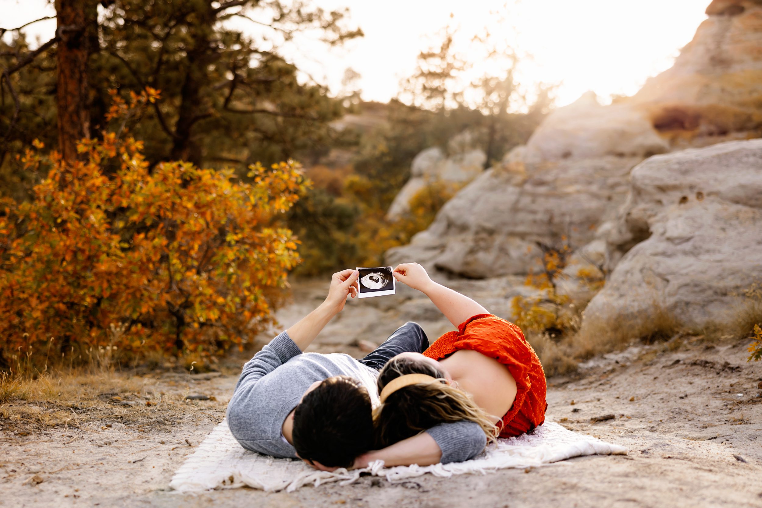 A couple lies on a blanket outdoors at sunset, holding up an ultrasound photo together with golden fall foliage and rocky terrain in the background