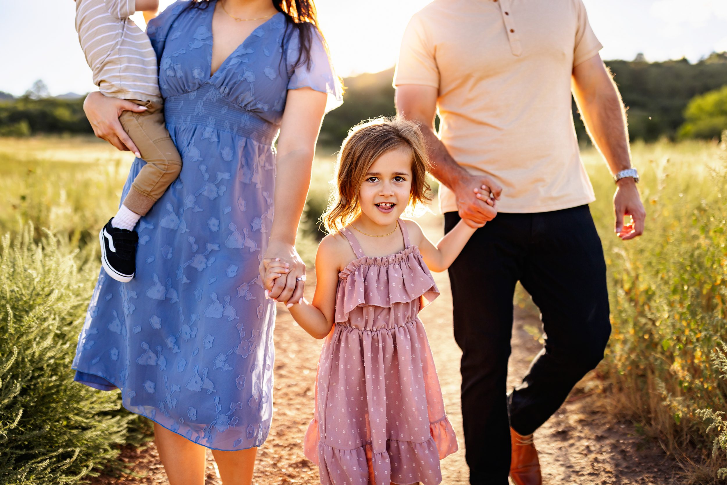Close-up of a young girl holding hands with her parents while walking through a grassy field during a summer family photo session.
