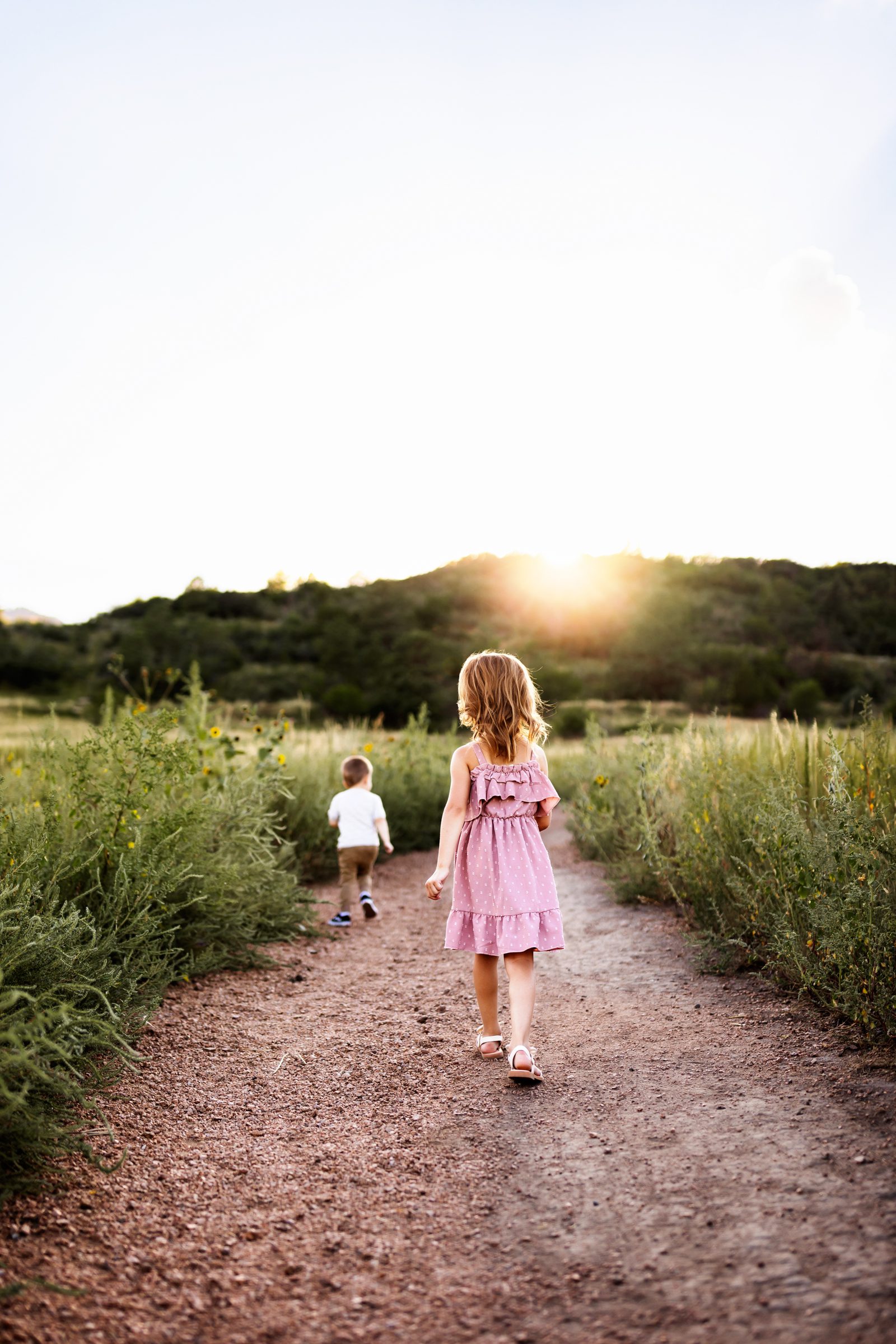 Young siblings walk ahead on a trail at sunset, surrounded by golden summer grass and mountain views.