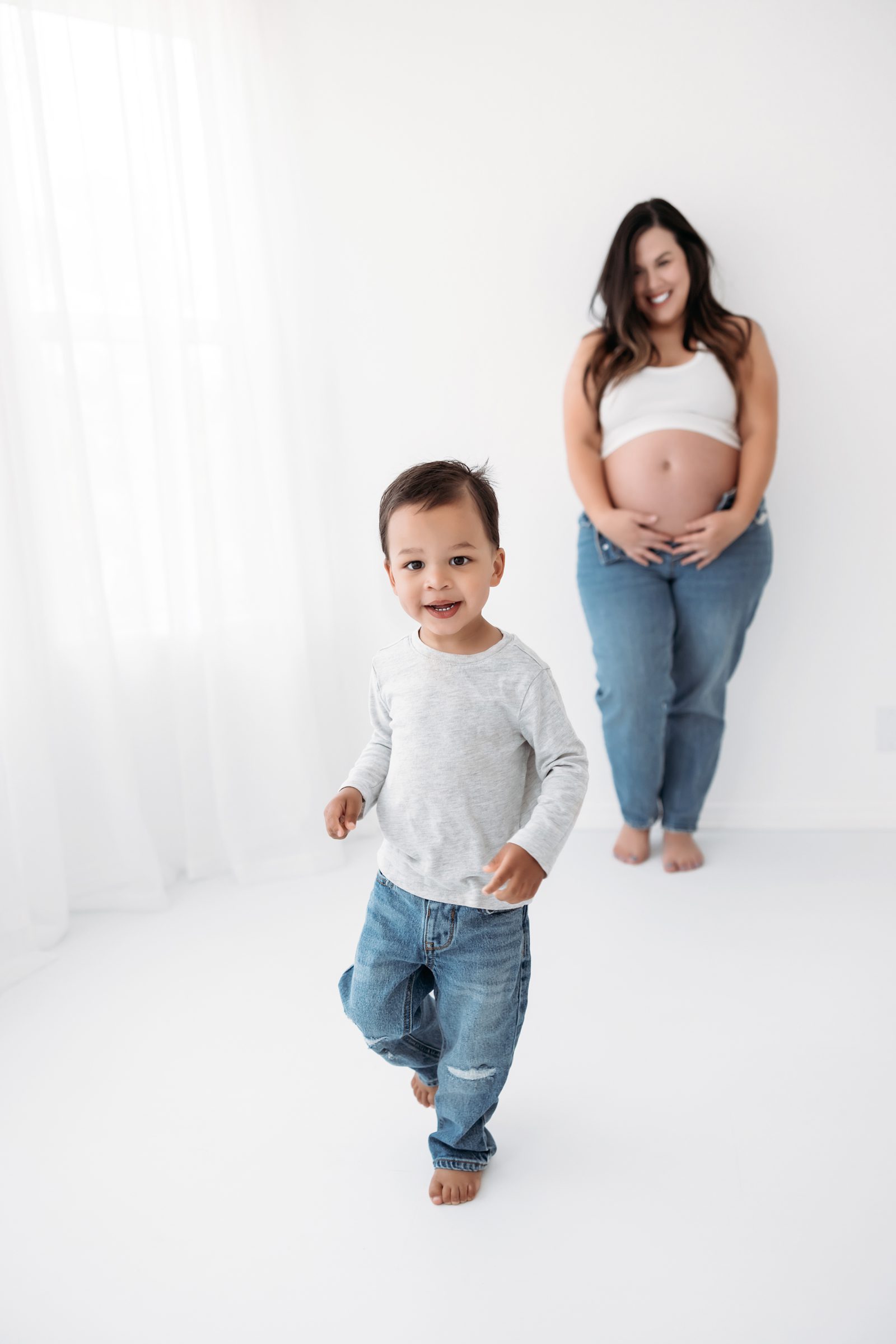 Toddler boy smiling at the camera while his mom stands in the distance smiling