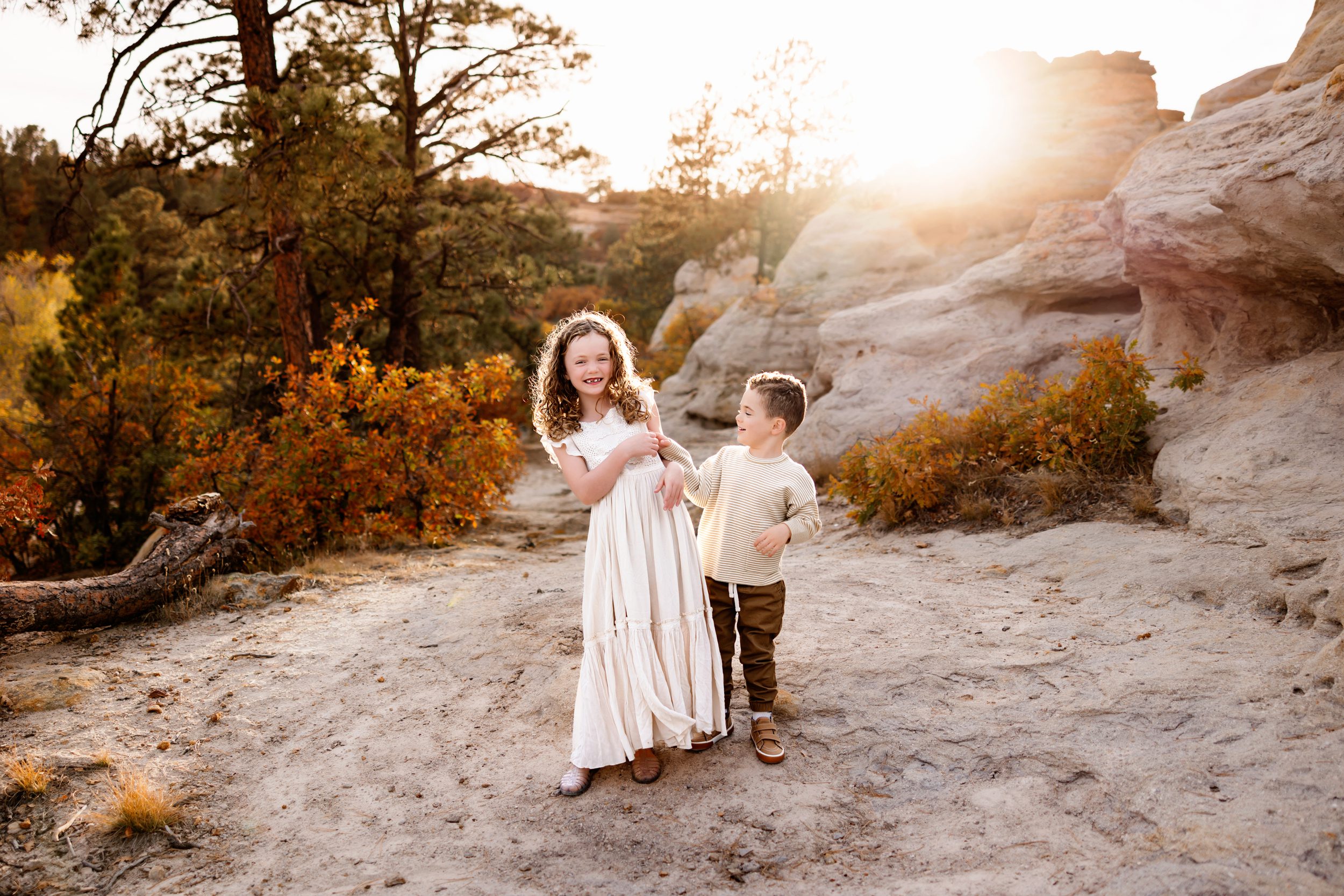 Two young siblings laugh together on a trail surrounded by fall leaves, glowing in the soft golden light of sunset.