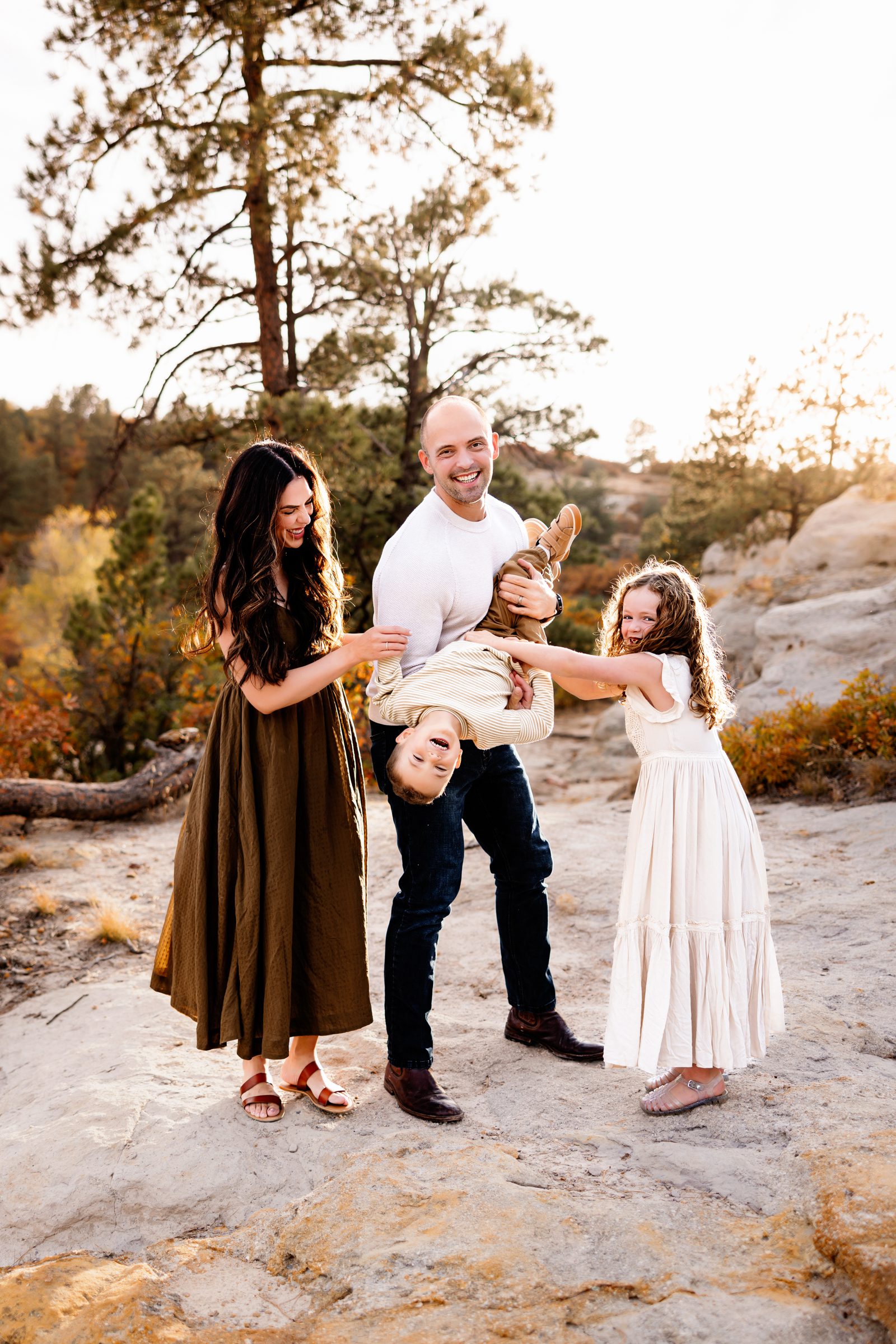 A playful family moment with parents and kids laughing as dad holds his son upside down while everyone smiles at each other in golden fall light.