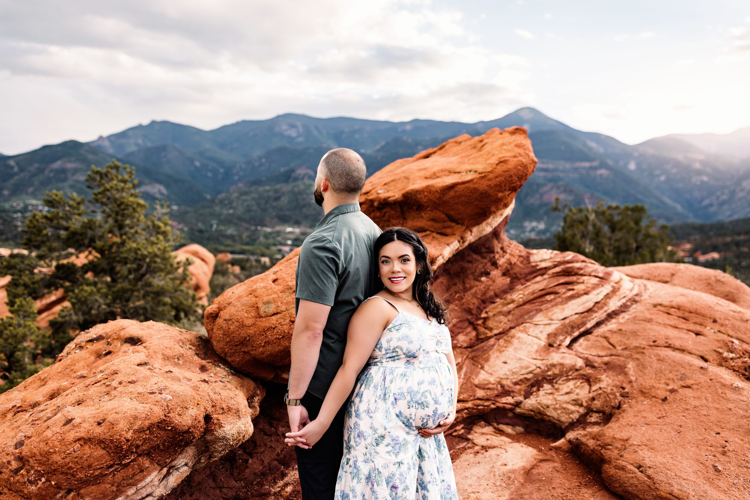Expecting couple standing back to back, holding hands among striking red rock formations with mountain peaks in the background.