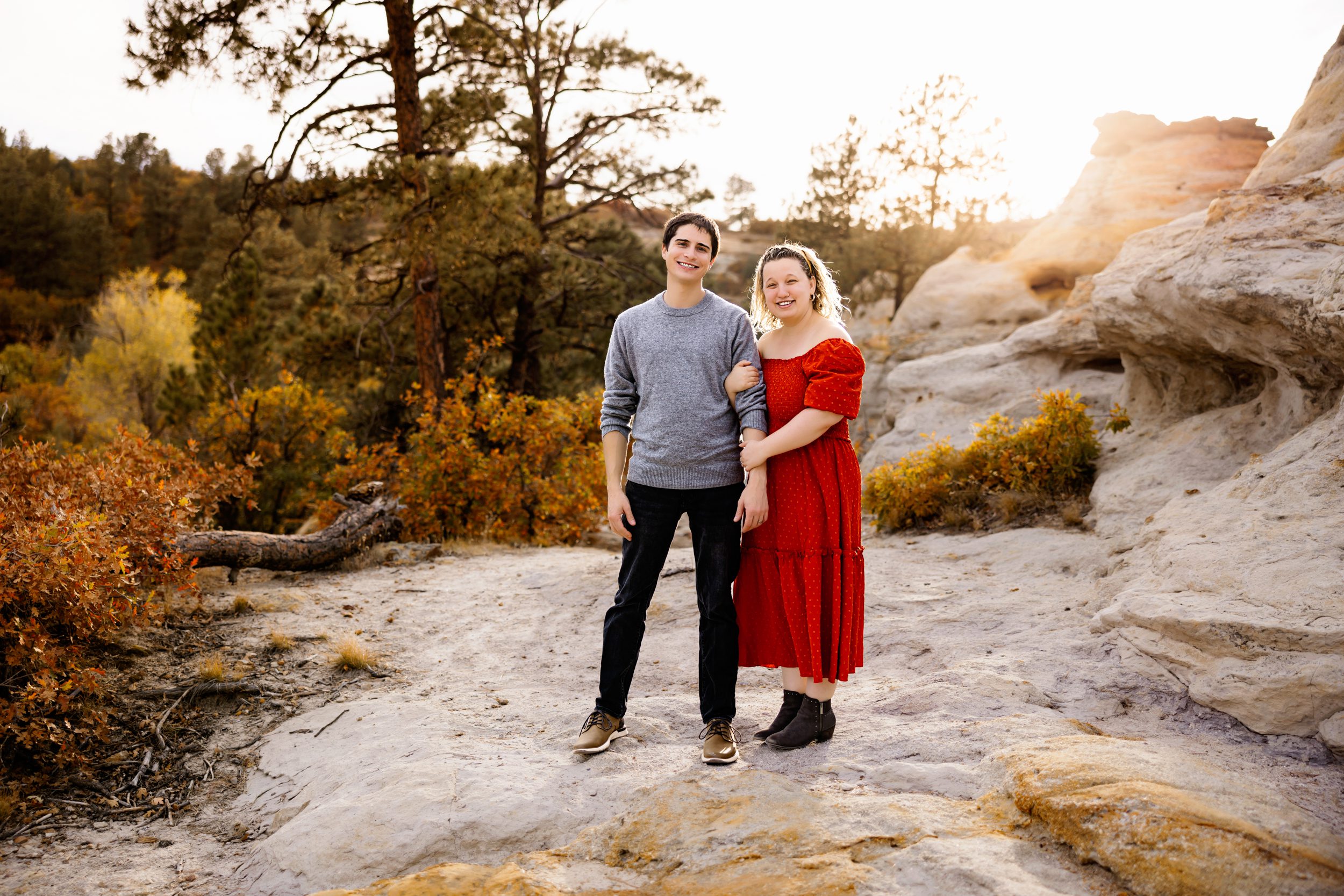 A couple smiles while standing arm in arm on a rocky path surrounded by golden autumn foliage and soft sunset light in Colorado Springs.