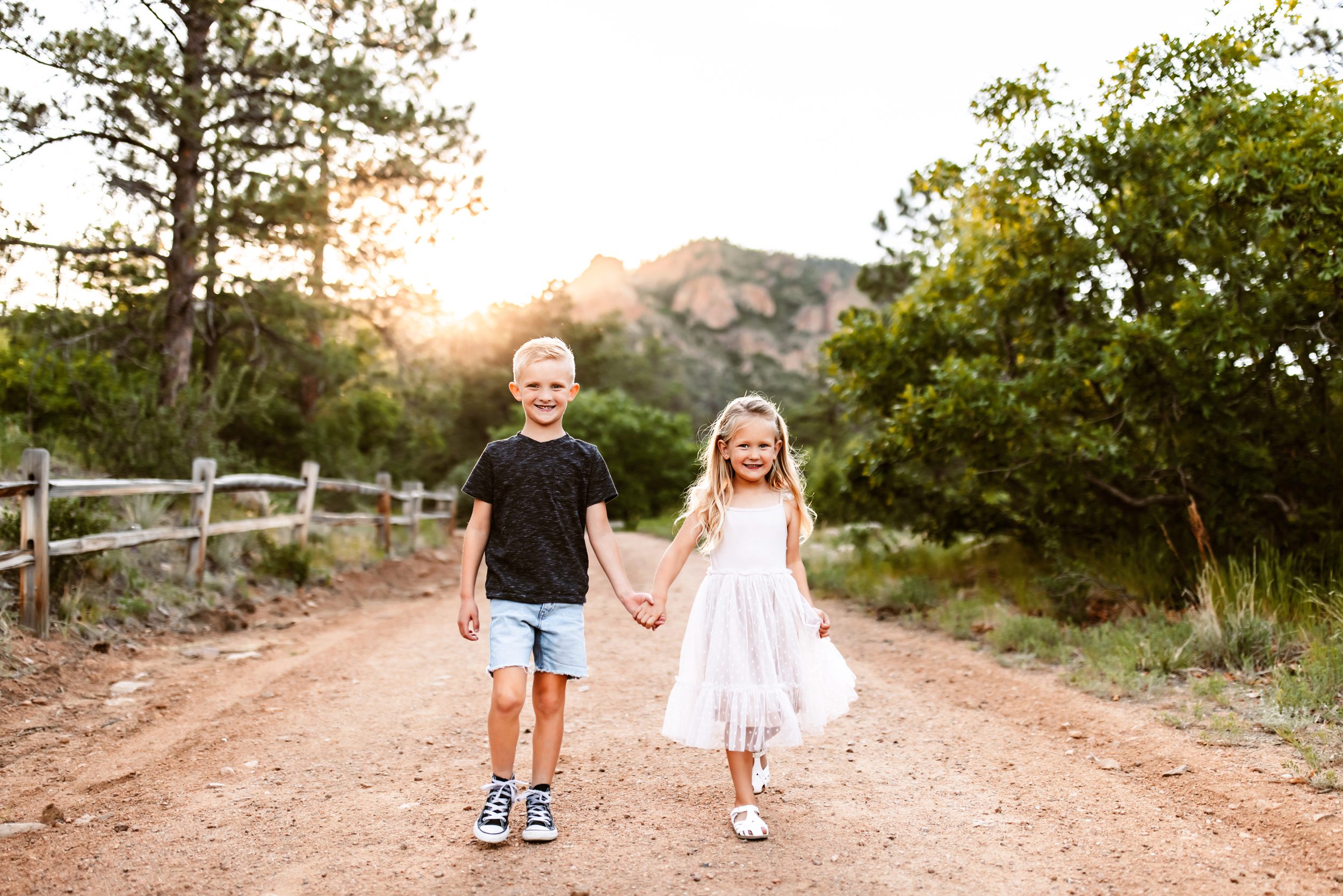 Smiling brother and sister holding hands on a dirt trail at sunset, surrounded by greenery and mountain views.