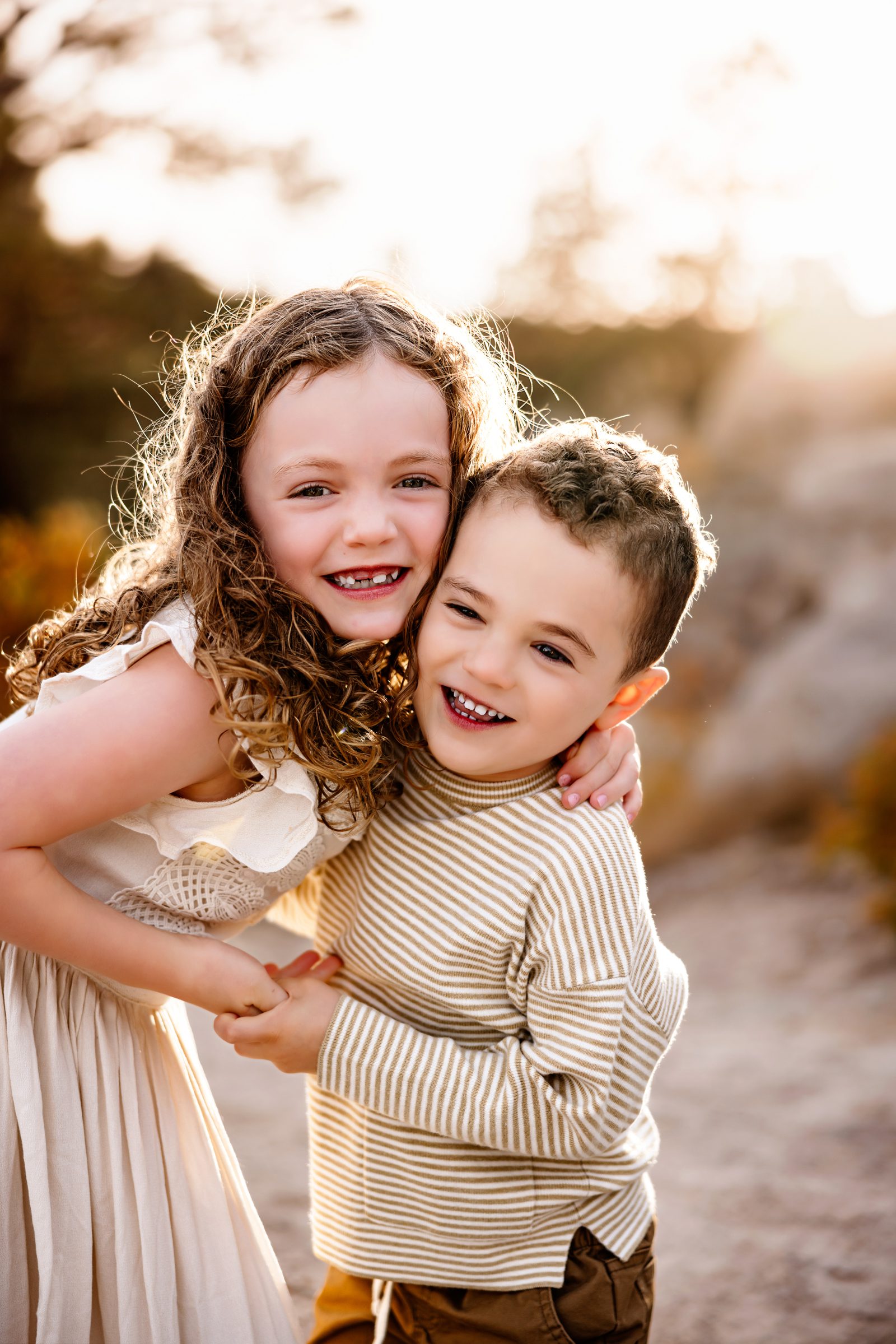 Big sister hugging her little brother close, both smiling with golden sunlight glowing around them during a playful outdoor moment.