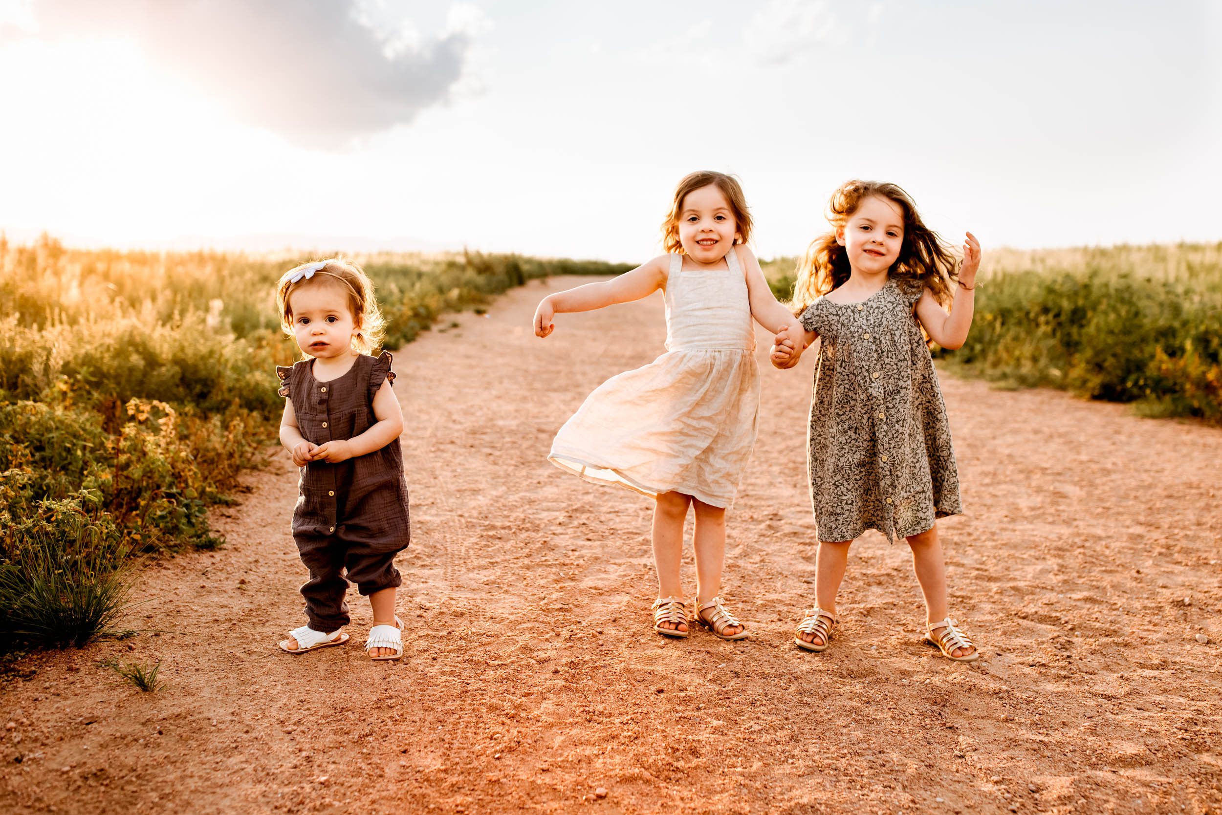 Three young sisters dancing and playing on a sunlit trail, with golden light glowing around them during a joyful summer evening.