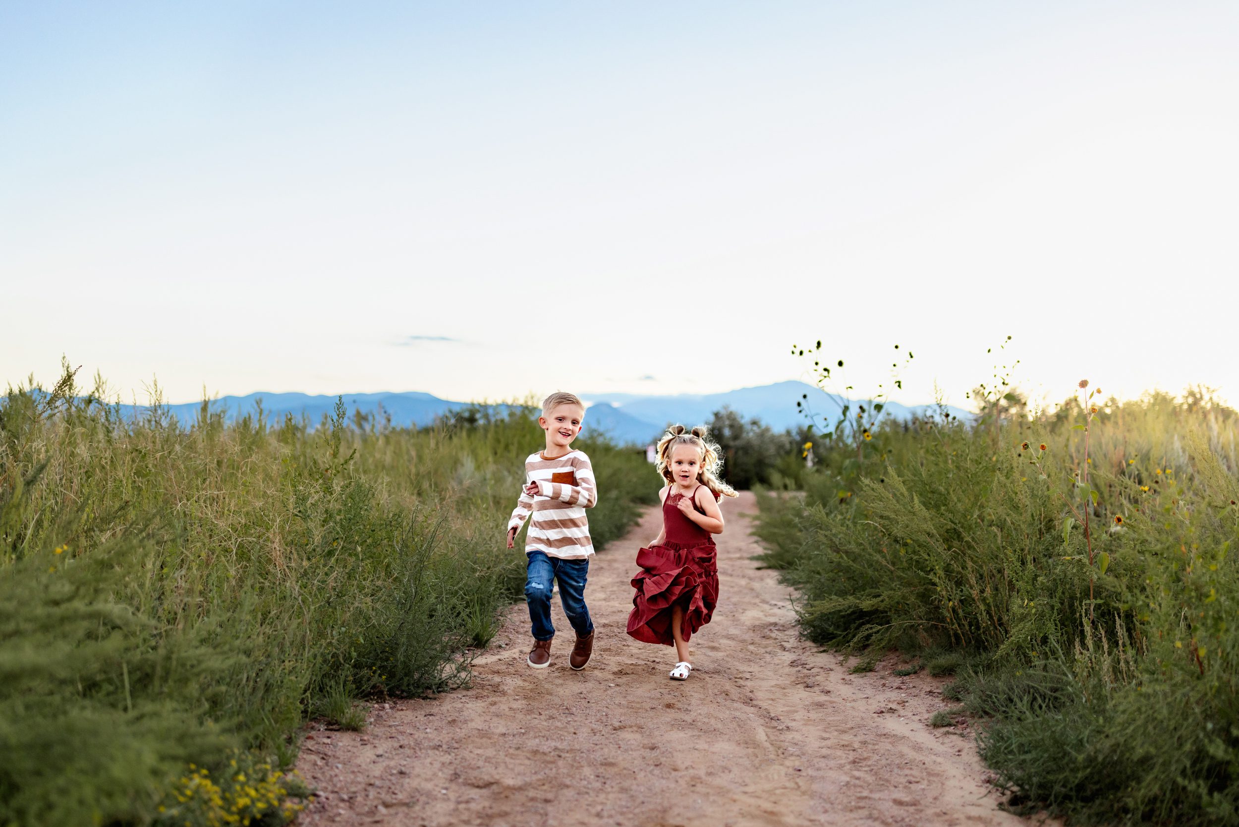 Brother and sister racing down a dirt path surrounded by wildflowers, captured by a sibling photos Colorado Springs photographer at golden hour.