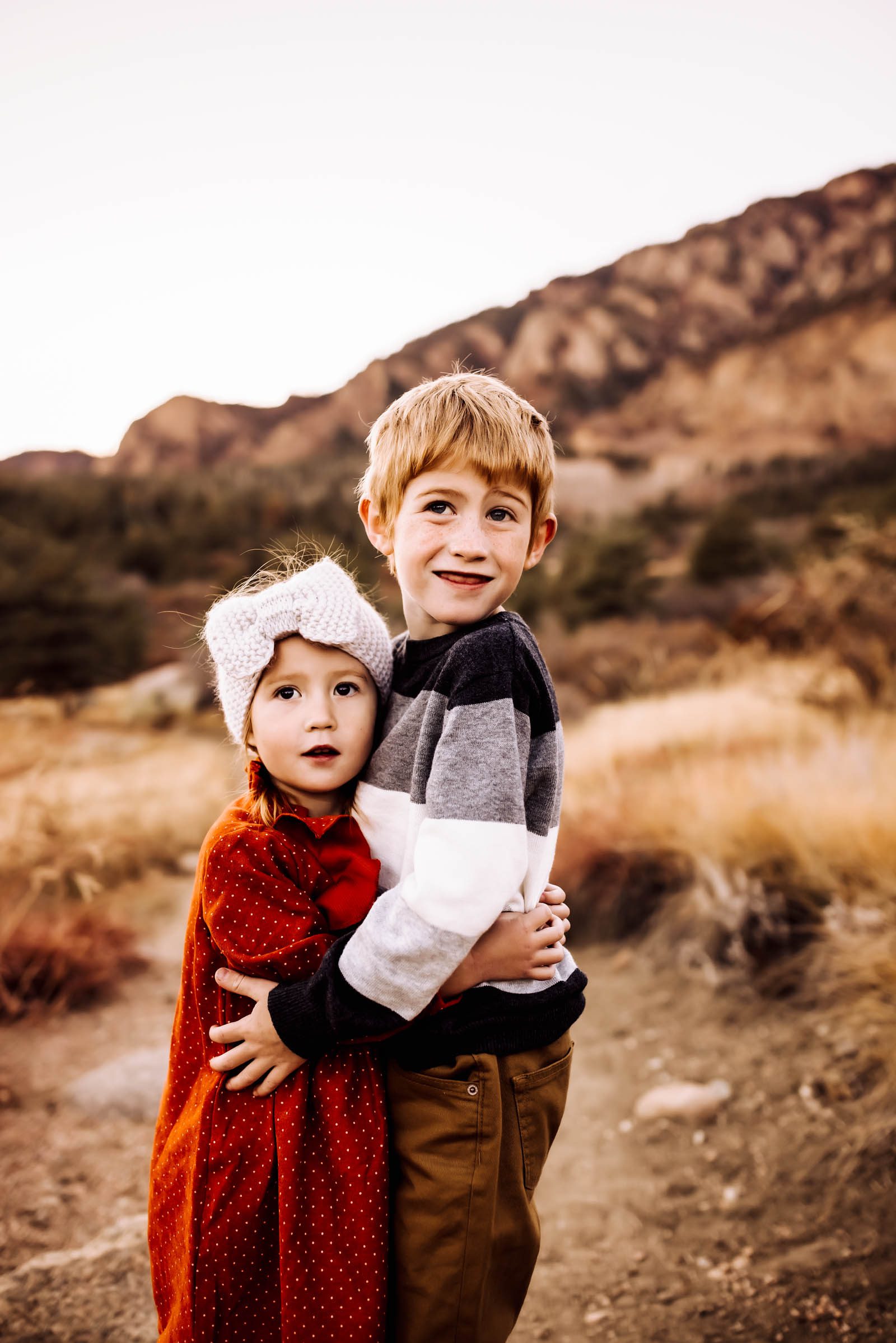Brother and sister hugging warmly in front of a mountain backdrop, dressed in cozy fall outfits with soft golden light around them.