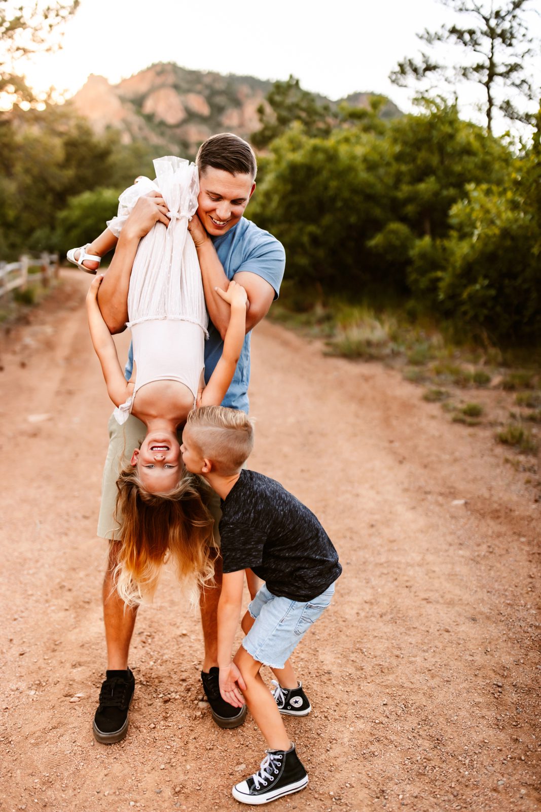 Dad playfully holds daughter upside-down while son kisses her cheek, sharing a sweet family moment